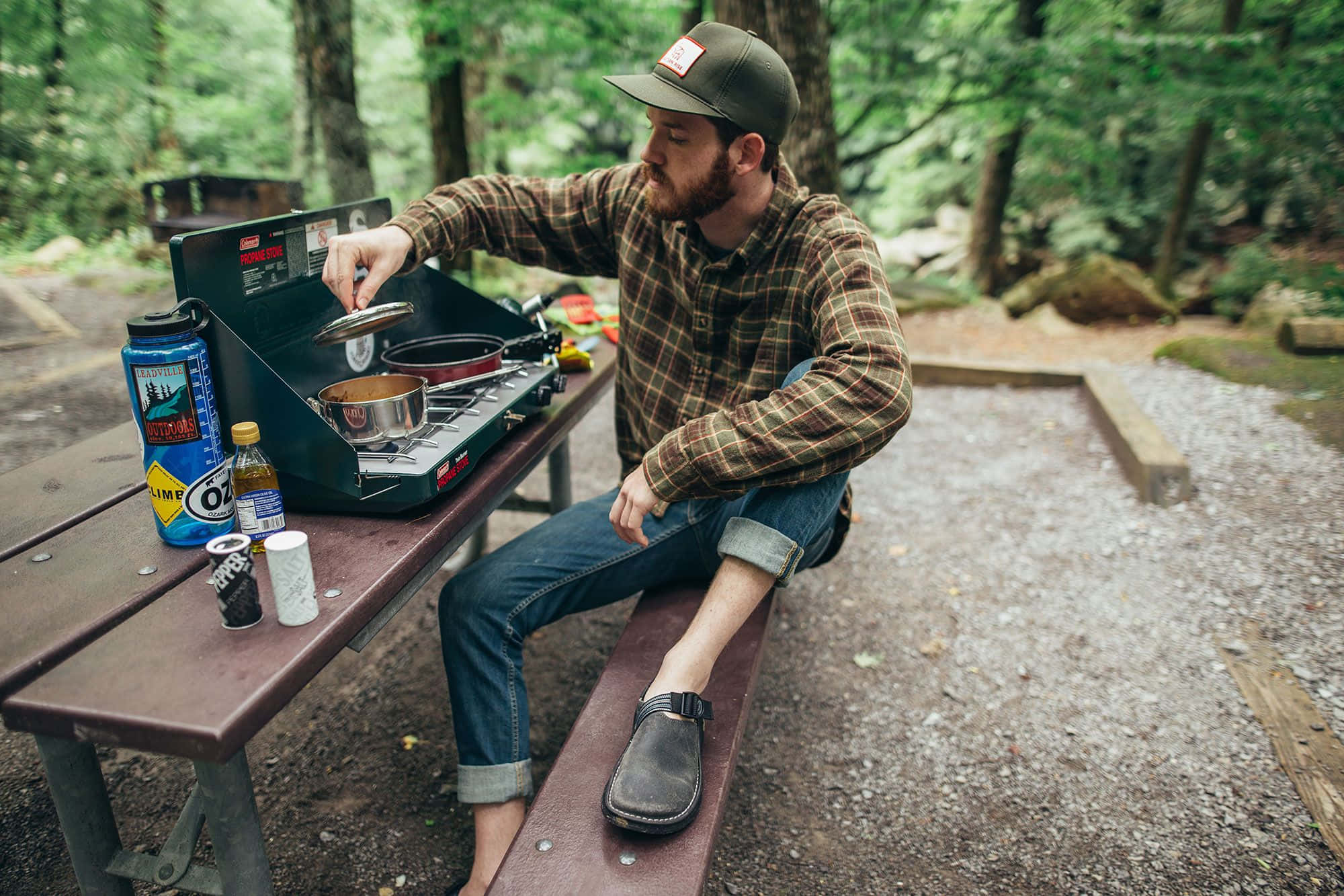Minimalist Camping 桌布 壁紙 — 一個留鬍子的男人穿著格子襯衫，在森林野餐桌旁用便攜式爐灶做飯，周圍是高聳的樹木。