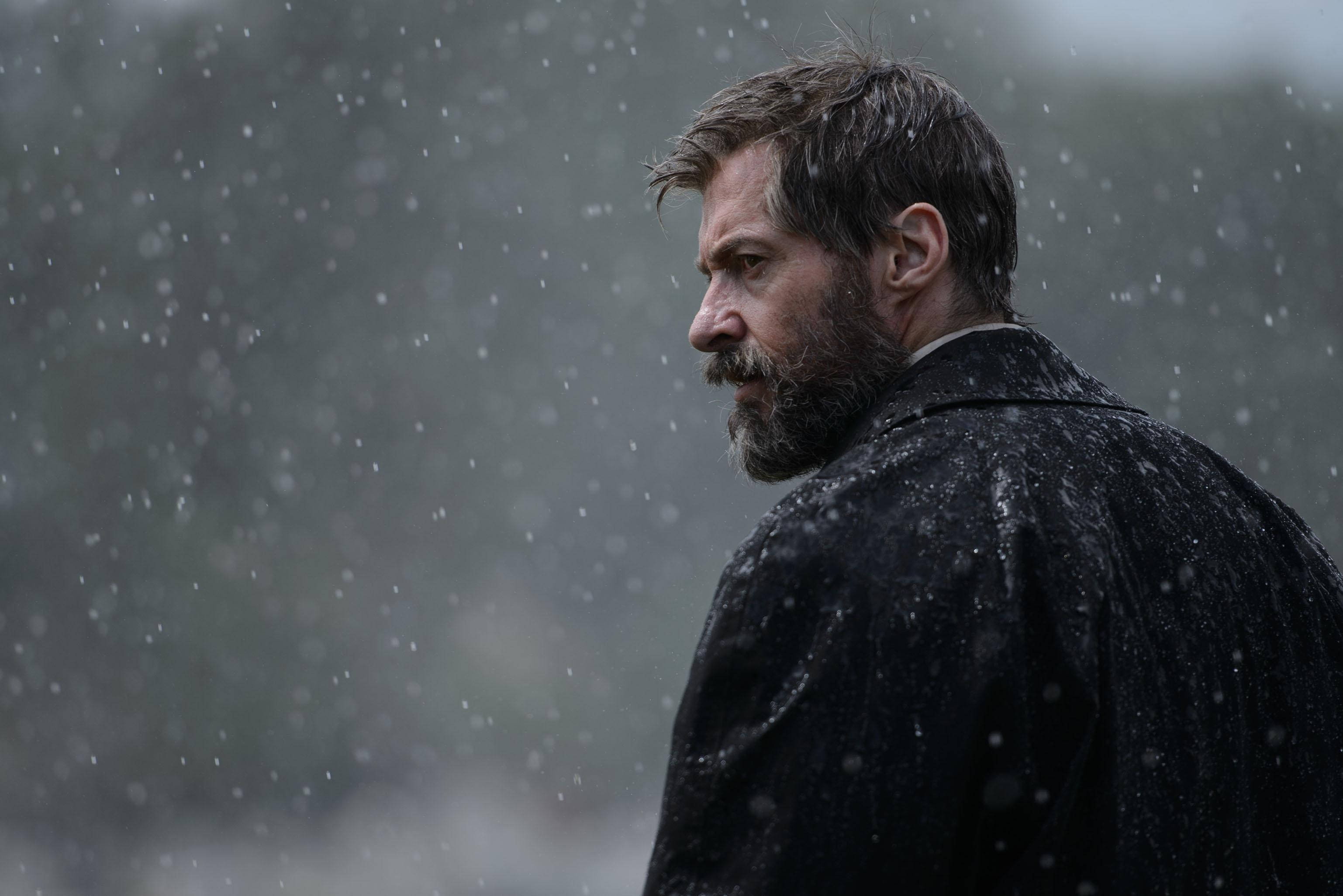 Hugh Jackman 桌布 壁紙 — 一個留鬍子的男人穿著深色外套，在傾盆大雨中站著，表情嚴肅，背景是模糊的灰色。