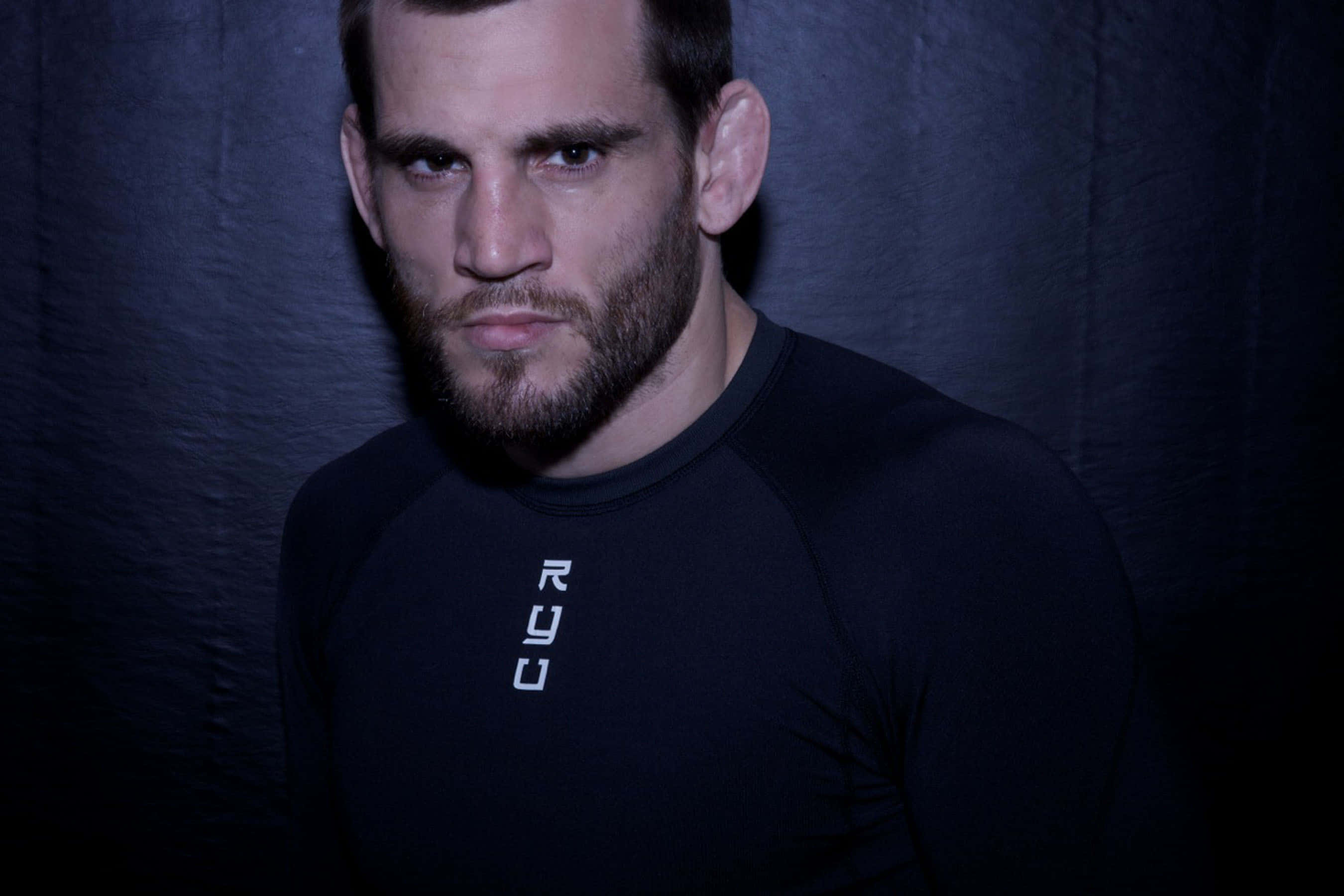 UFC 桌布 壁紙 — 一個留鬍子的男人穿著黑色運動衫，在戲劇性的燈光下，對著深色紋理背景強烈凝視。