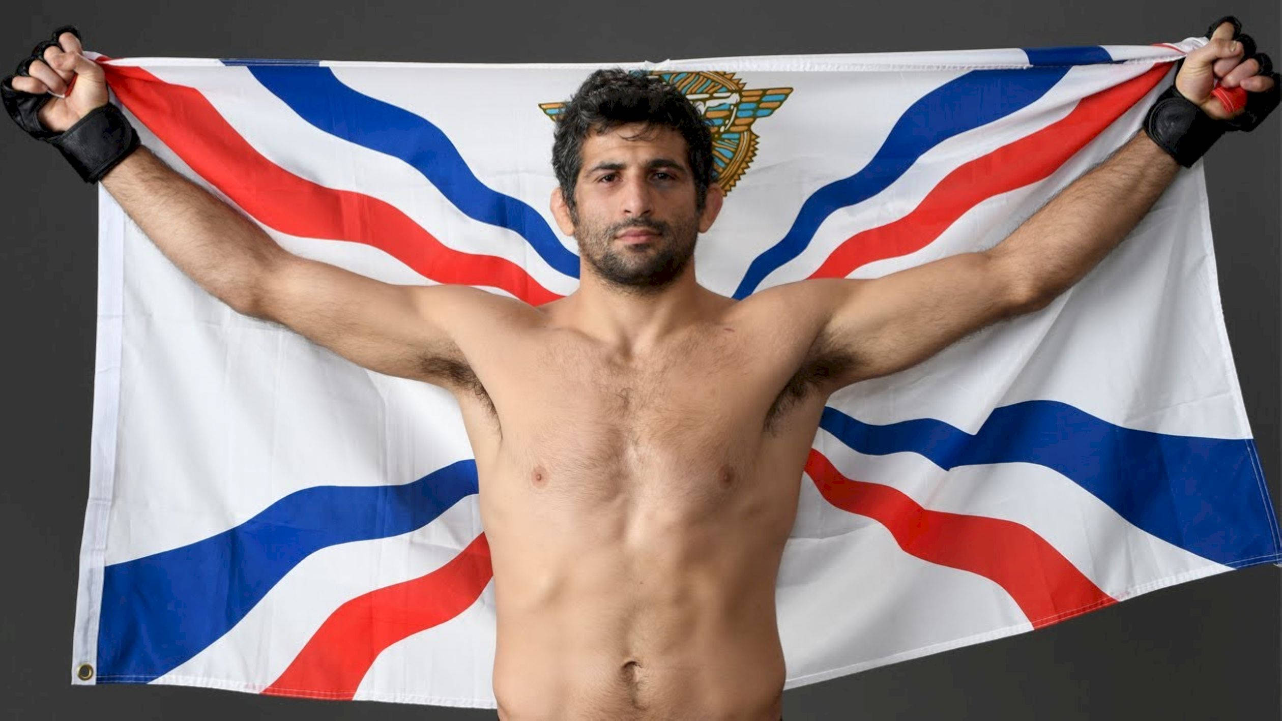 UFC 桌布 壁紙 — 一個留鬍子的肌肉男在深色背景上，將一面帶有紋章的紅白藍旗幟高舉過頭。