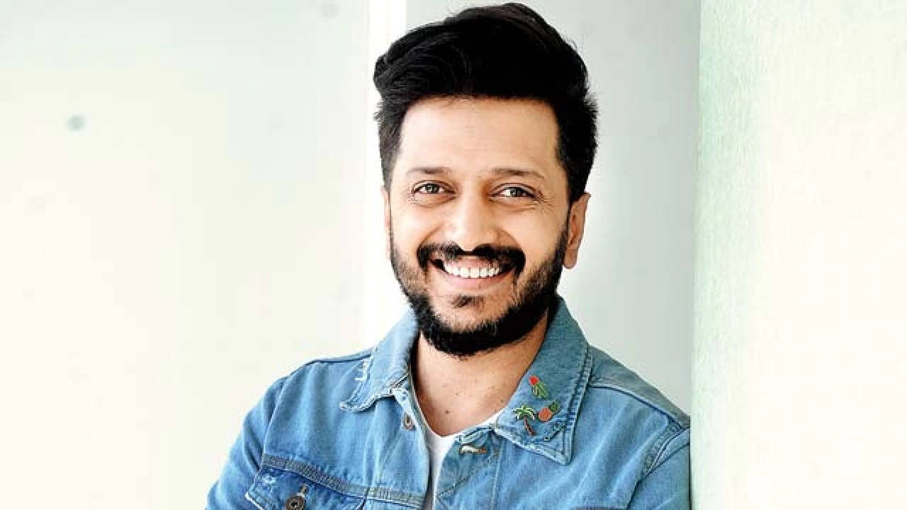 riteish deshmukh 桌布 壁紙 — 一個留鬍子的開朗男人，穿著牛仔外套，靠著淺色牆壁，溫暖地微笑。