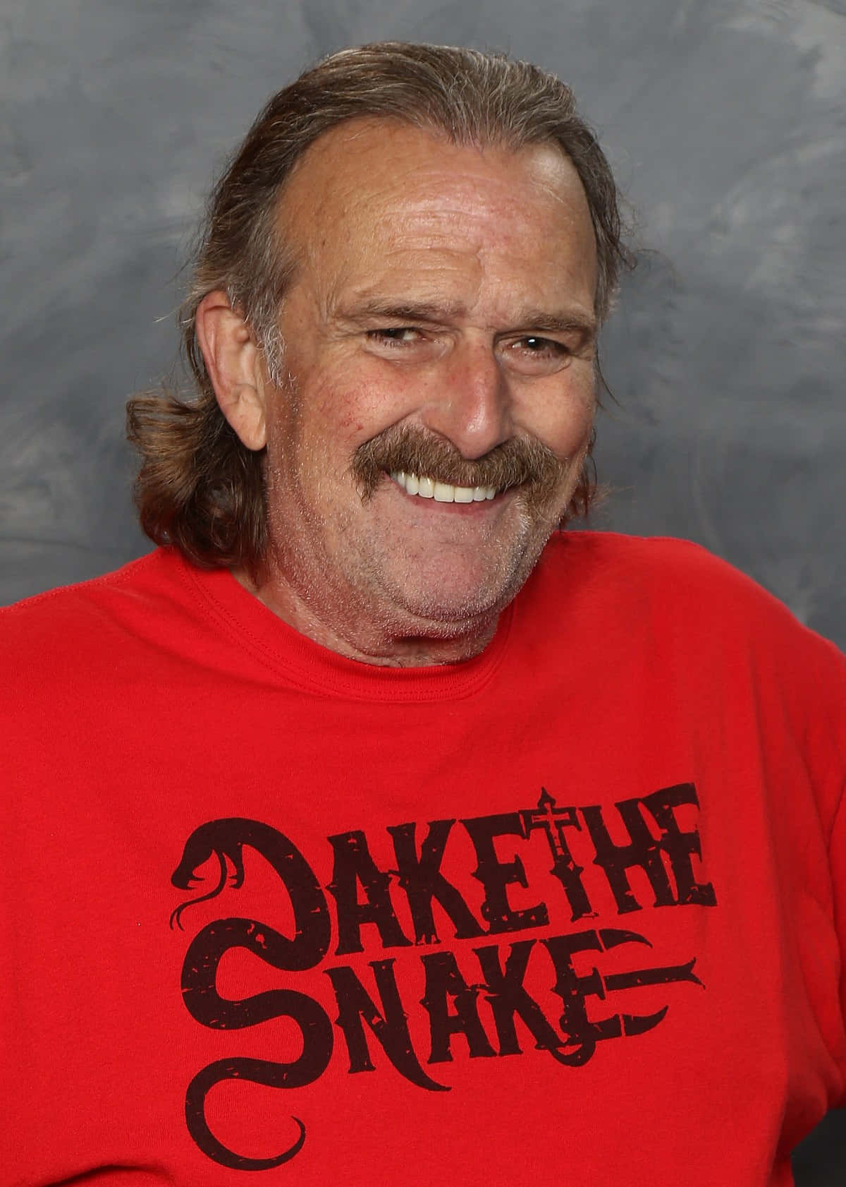 摔角 桌布 壁紙 — 一個留鬍子長髮的開朗男人，穿著印有 'DAKE THE SNAKE' 圖案的紅色T恤微笑。