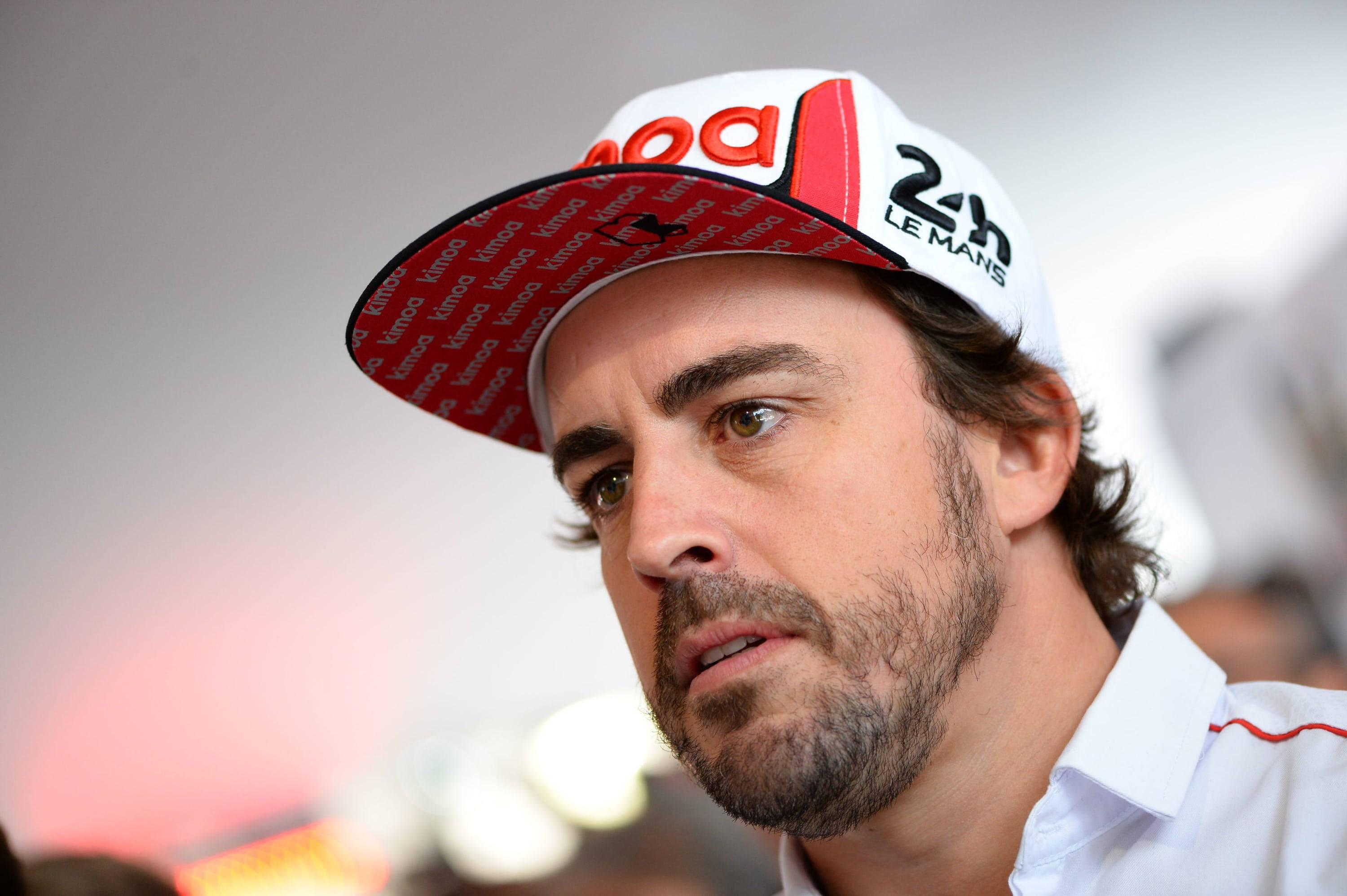 Fernando Alonso 桌布 壁紙 — 一個留鬍鬚、專注的男人，戴著一頂帶有 24h Le Mans 品牌標誌的白色紅色帽子，背景柔和模糊。
