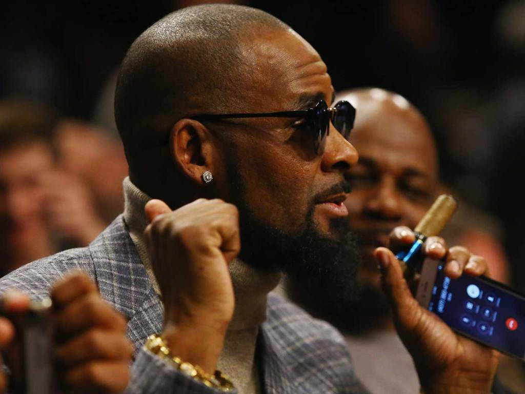 r kelly 桌布 壁紙 — 一個留鬍鬚、戴太陽眼鏡的男人在昏暗的人群中熱情地做手勢，同時拿著手機。