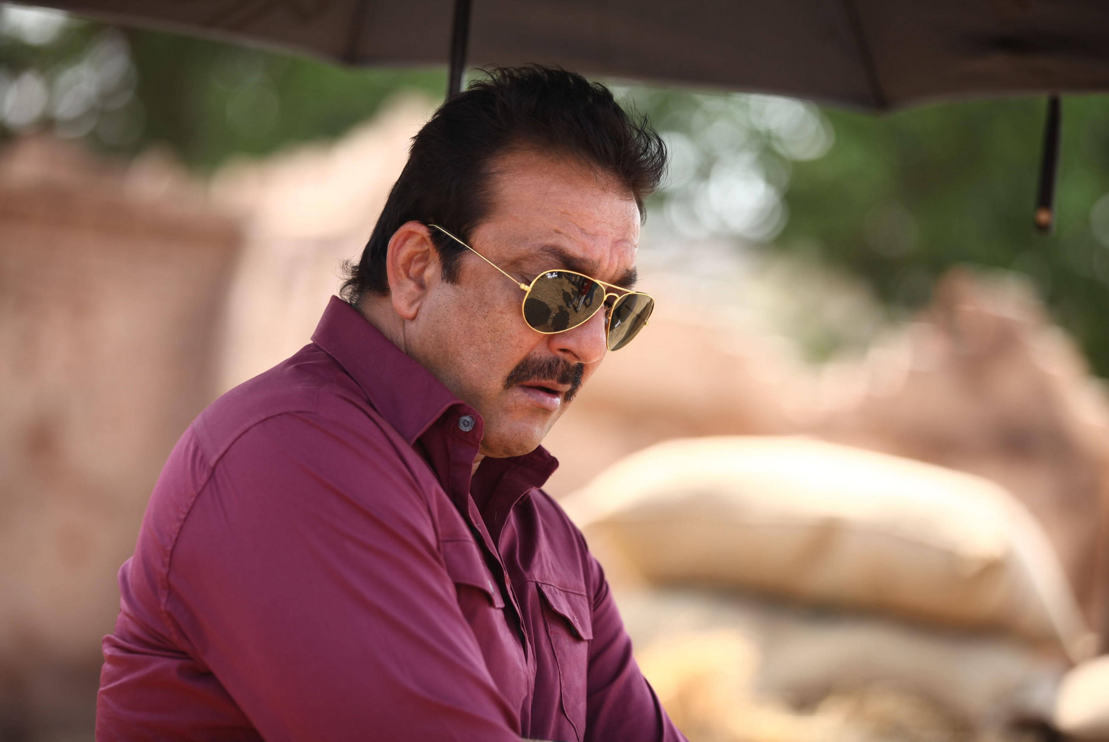 Sanjay 桌布 壁紙 — 一個留鬍鬚、戴金色太陽眼鏡的男人，穿著栗色襯衫，坐在雨傘下，背景是模糊的泥土色。