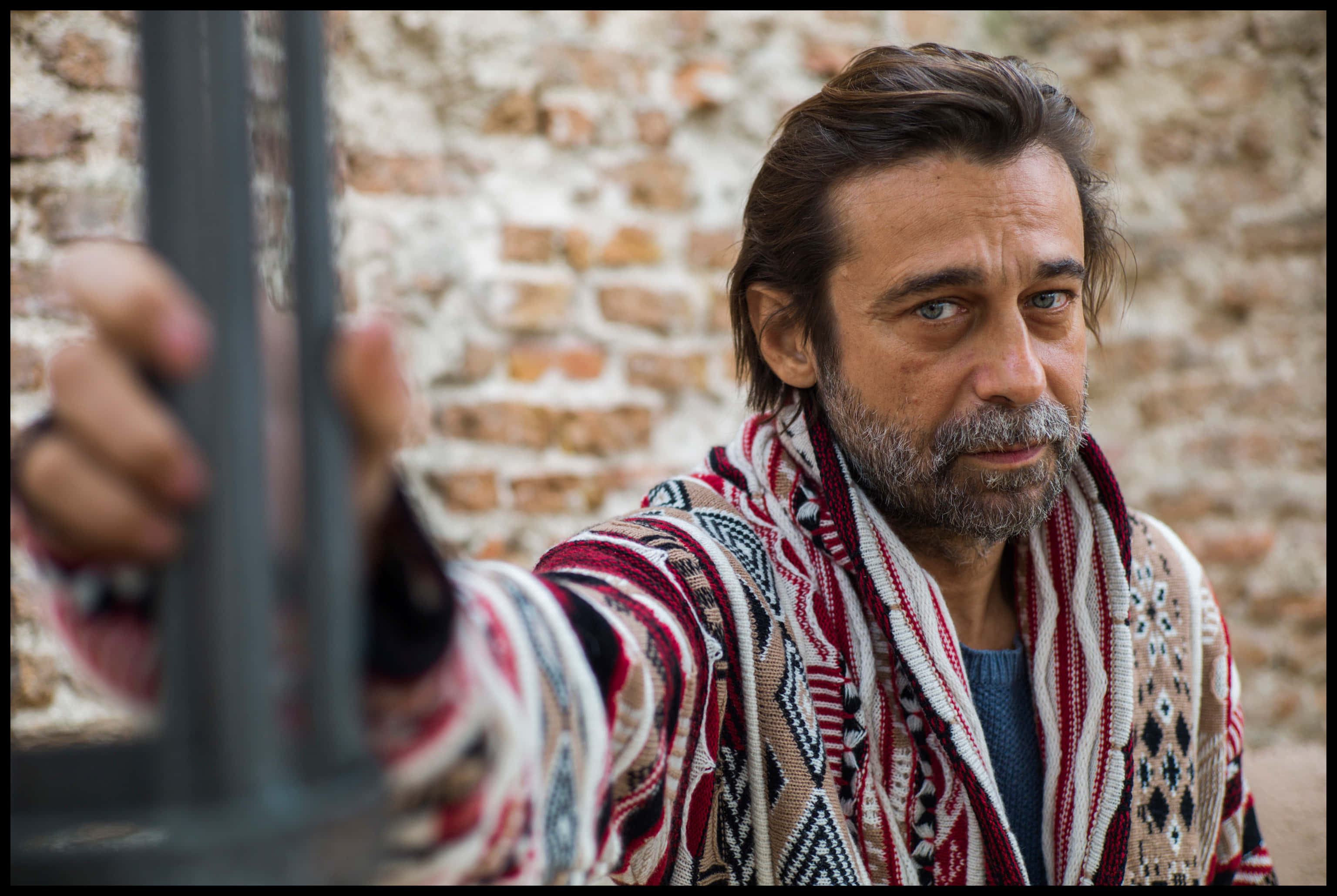 Jordi Mollà 桌布 壁紙 — 一個留鬍鬚、披著圖案披肩的男人，抓著金屬棒靠在風化的磚牆上，目光專注地直視前方。