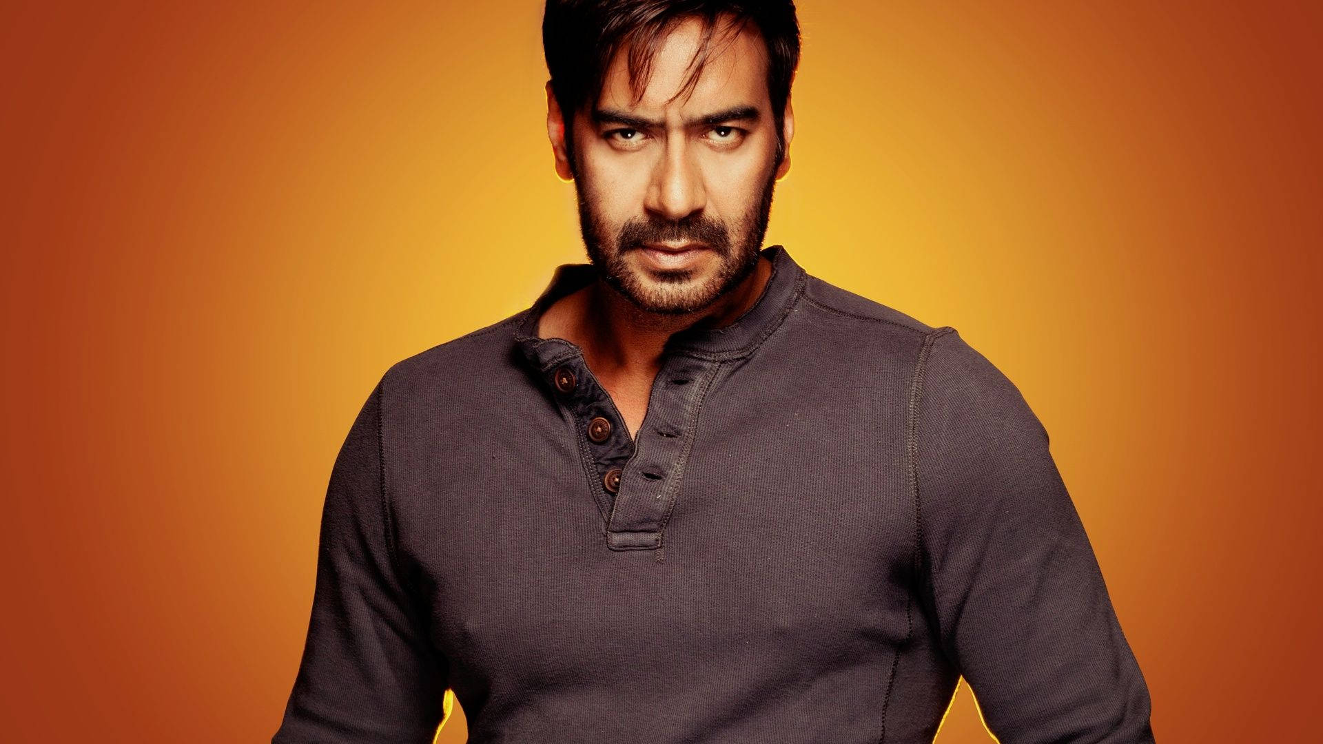 ajay devgn 桌布 壁紙 — 一個留鬍鬚、眼神銳利的男人，穿著深色亨利衫，背景是溫暖的橙色漸層。