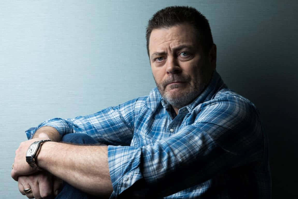 nick offerman 桌布 壁紙 — 一個留鬍鬚、眼神銳利的男人，身穿藍色格紋襯衫，向前傾身坐在柔和的灰色背景前。