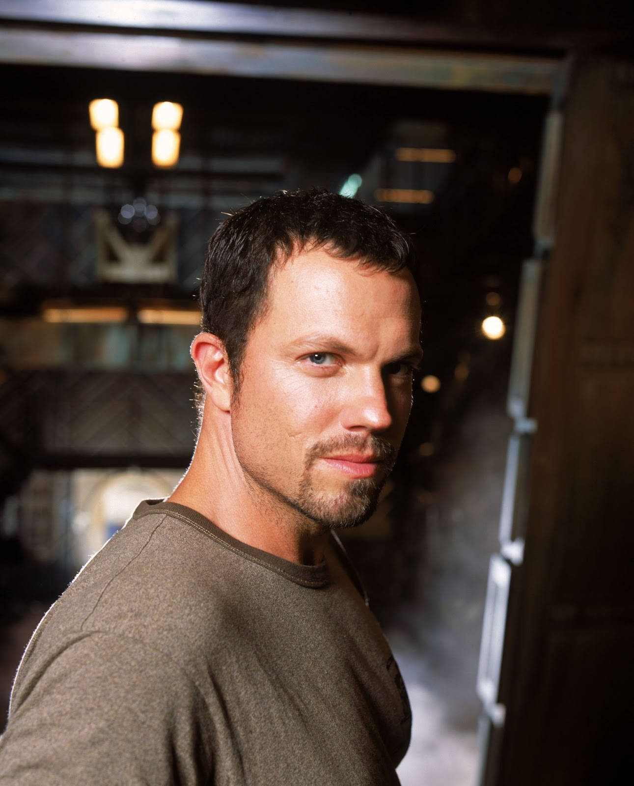adam baldwin 桌布 壁紙 — 一個留鬍鬚、眼神銳利的男人在昏暗的工業環境中回頭看，背景有溫暖的散景燈光。