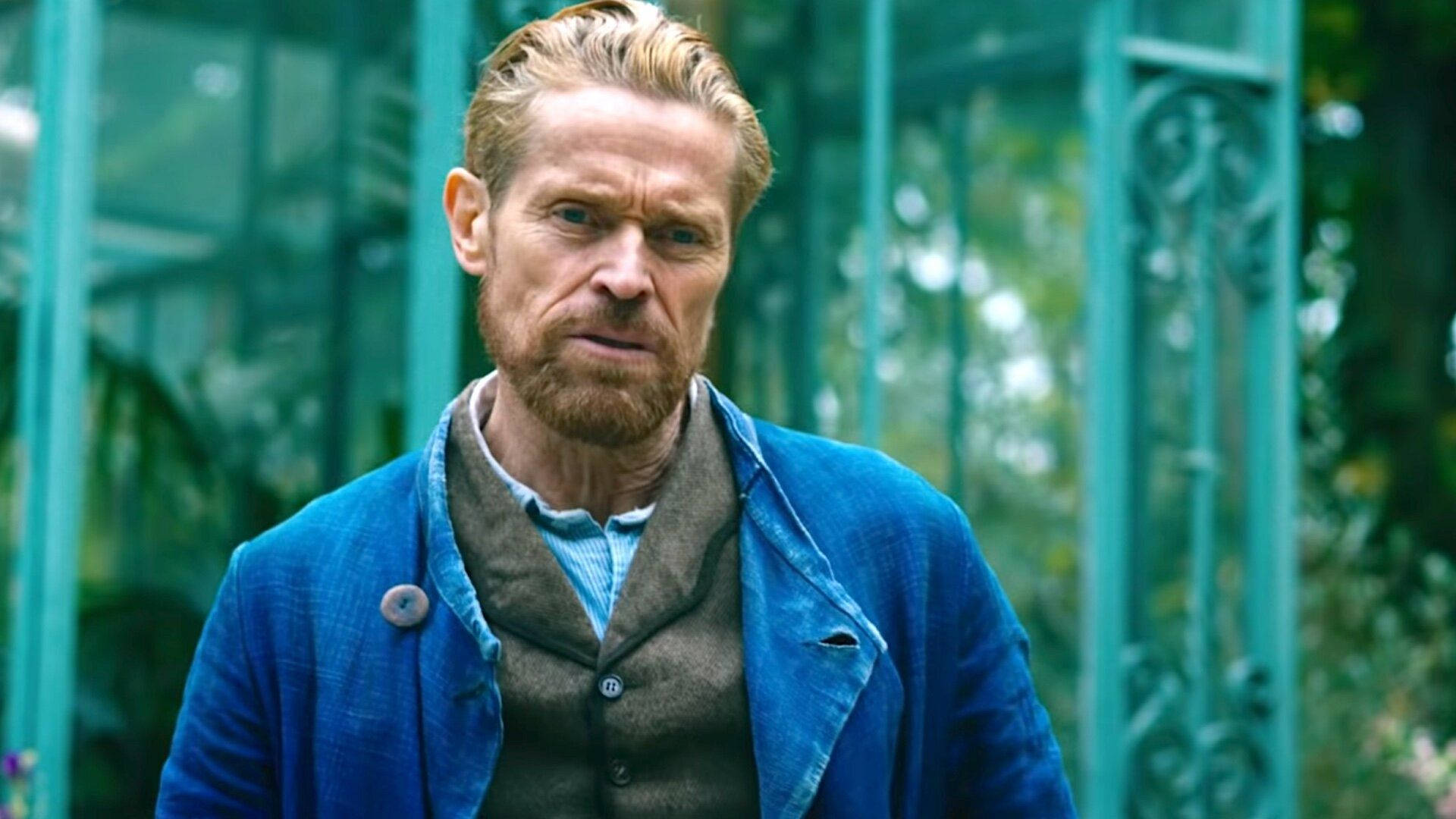 Willem Dafoe 桌布 壁紙 — 一個留鬍鬚、眼神銳利的男人站在一個被綠色植物包圍的藍綠色金屬花園門前。