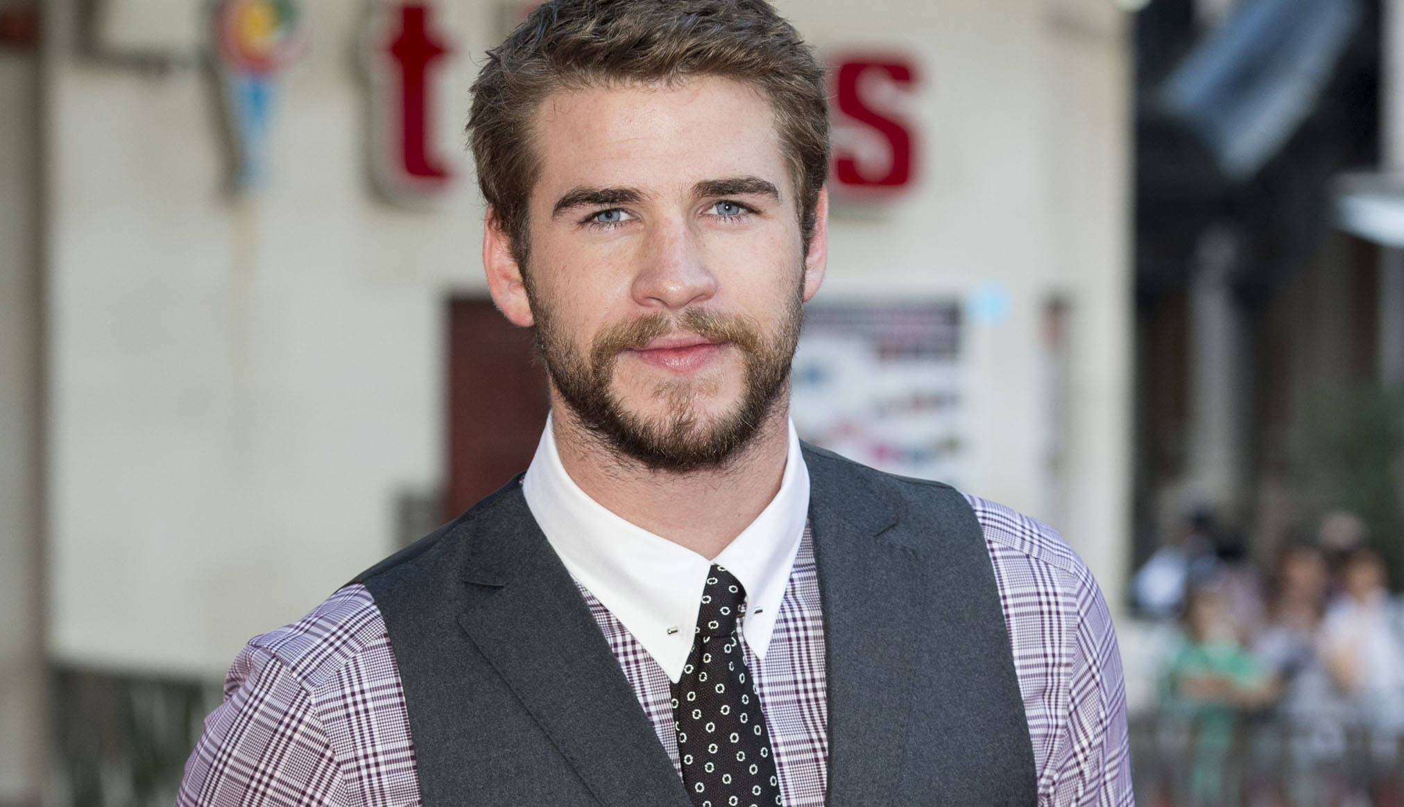 liam 桌布 壁紙 — 一個留鬍鬚、藍眼睛的男人，穿著格紋襯衫背心和圓點領帶，背景是模糊的城市景觀。