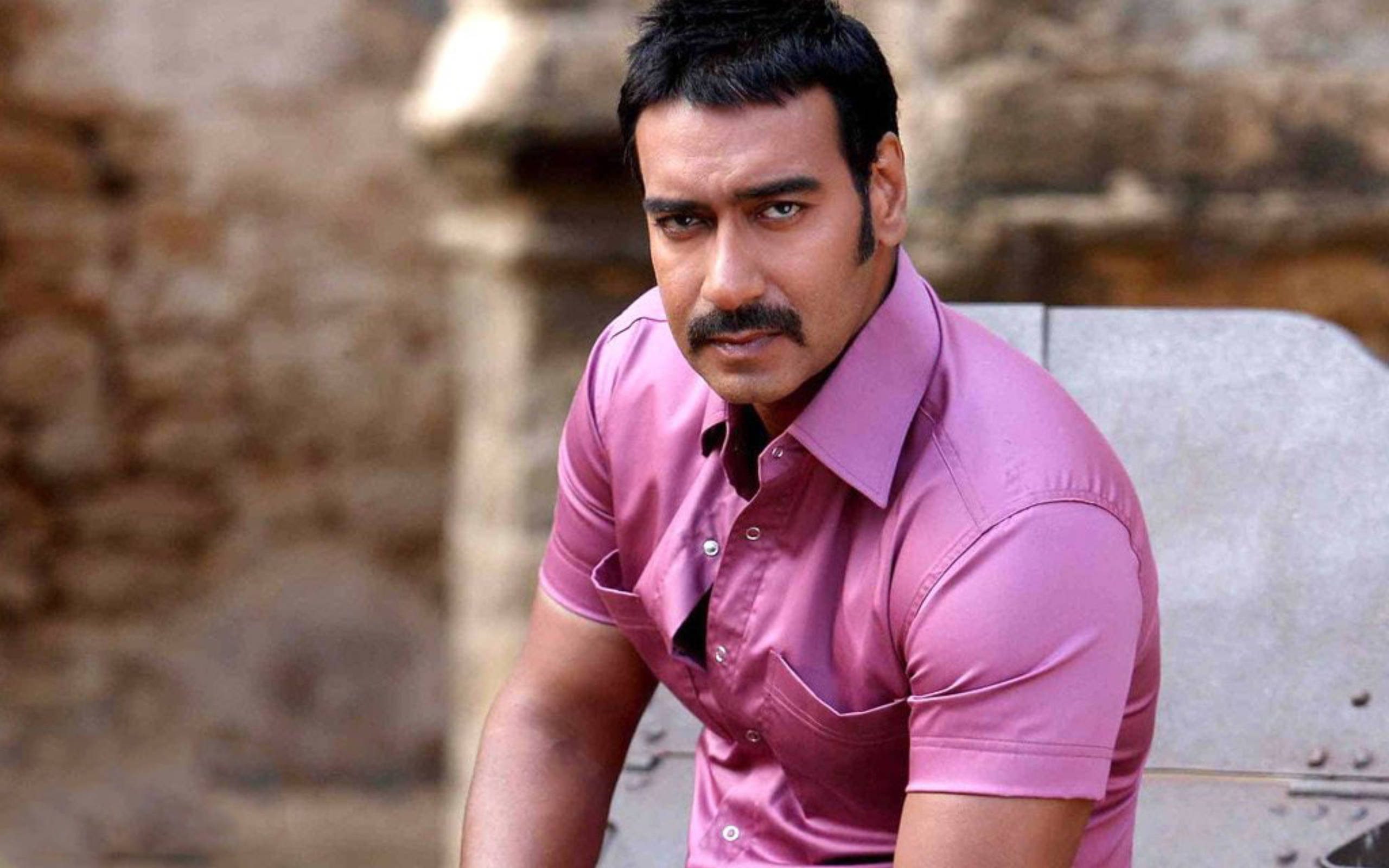 ajay devgn 桌布 壁紙 — 一個留鬍鬚、身穿粉紅色襯衫的男人，帶著嚴肅的眼神靠在石結構上。