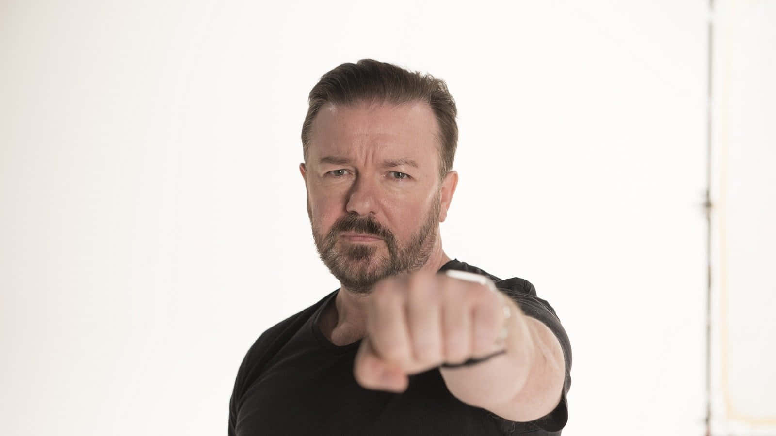 ricky gervais 桌布 壁紙 — 一個留鬍鬚、身穿黑色襯衫的男人，在純白背景前直接指向觀眾。