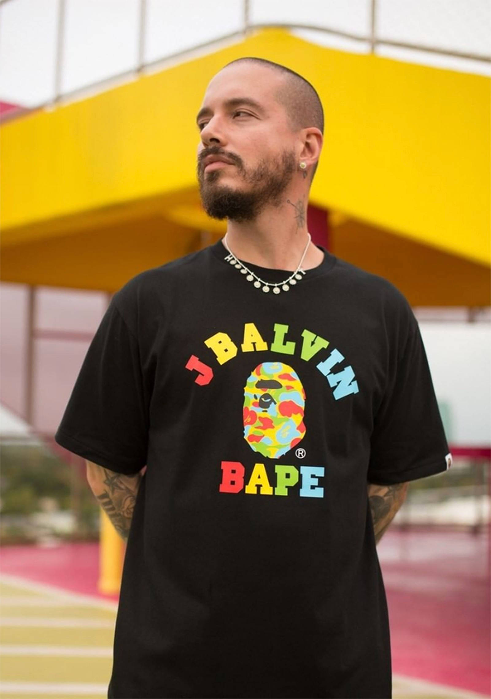 J 桌布 壁紙 — 一個留鬍鬚和有刺青的男人，穿著一件黑色T恤，上面有色彩繽紛的Bape標誌，背景是明亮的黃色建築。