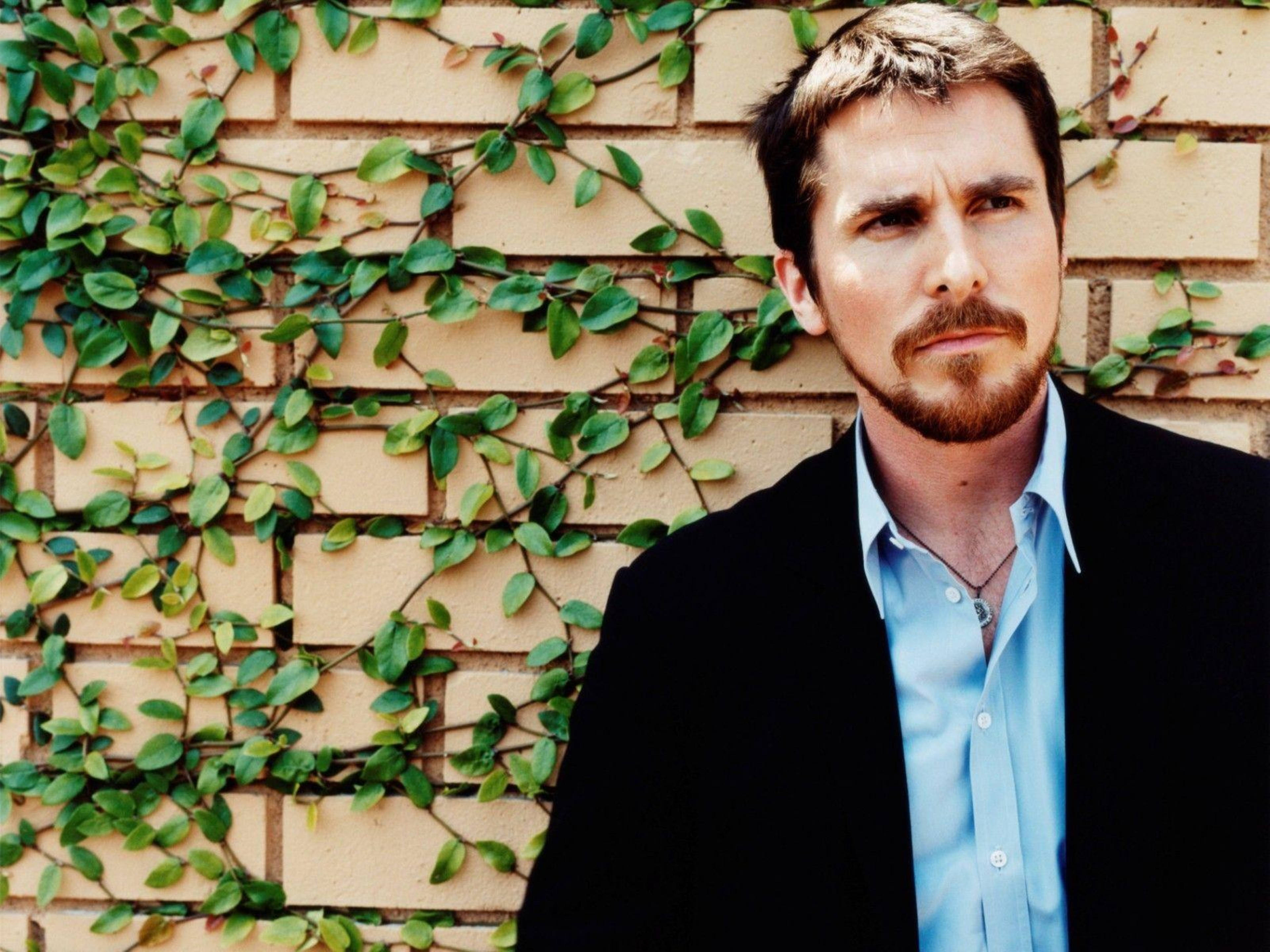 Christian Bale 桌布 壁紙 — 一個留鬍鬚和鬍子的男人，身穿黑色西裝外套和藍色襯衫，靠在一面部分被綠色常春藤覆蓋的磚牆上。