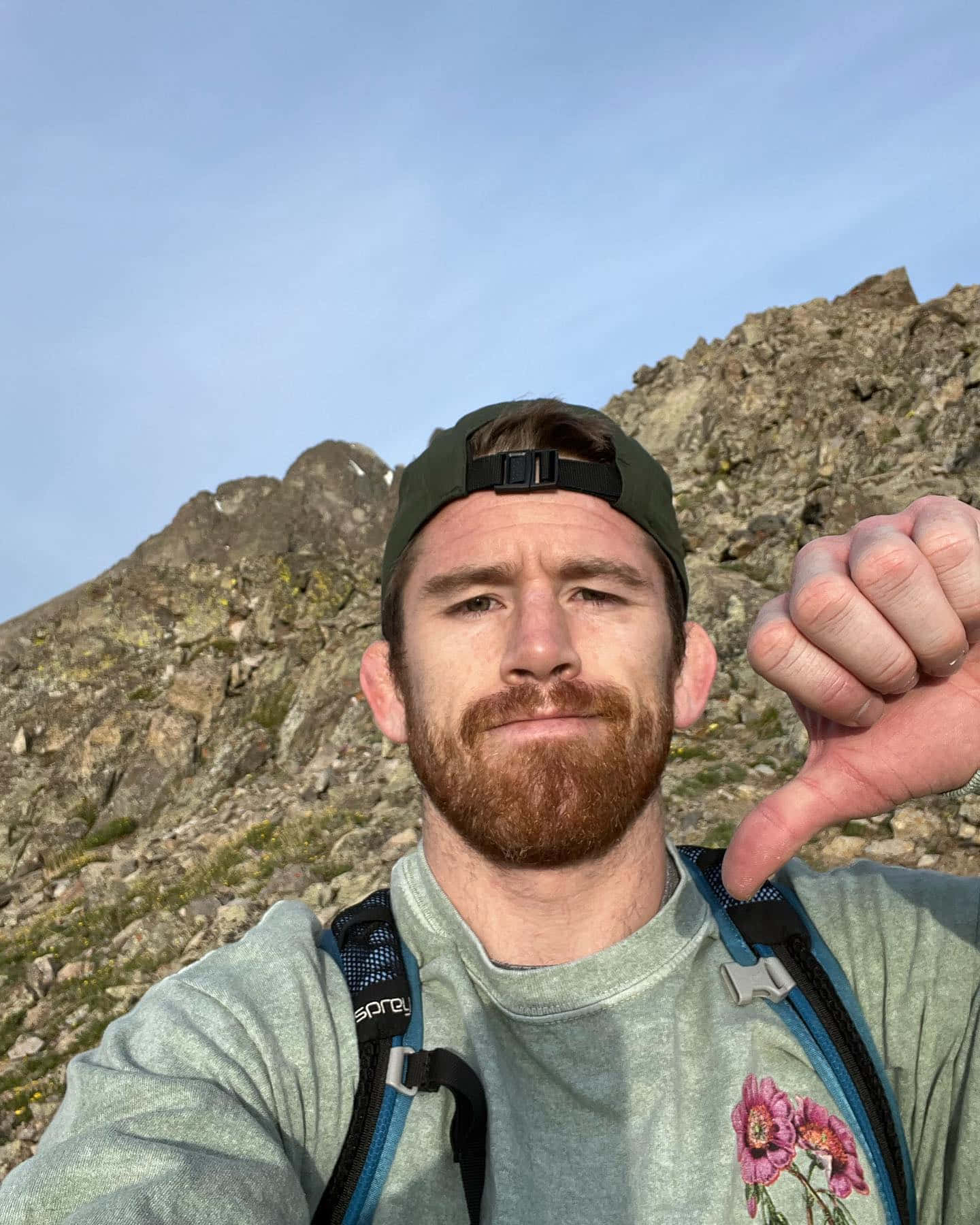 Hiking 桌布 壁紙 — 一個留鬍鬚戴帽子的男人在晴朗藍天下，以岩石山脈為背景徒步旅行時豎起拇指向下。