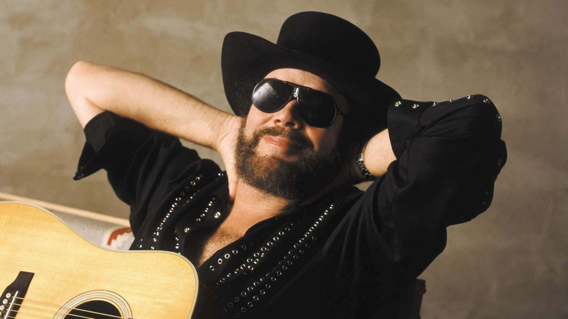 Hank Williams Jr 桌布 壁紙 — 一個留鬍鬚的男人，戴著黑色牛仔帽和太陽眼鏡，雙手放在腦後，向後靠著，拿著一把木吉他。
