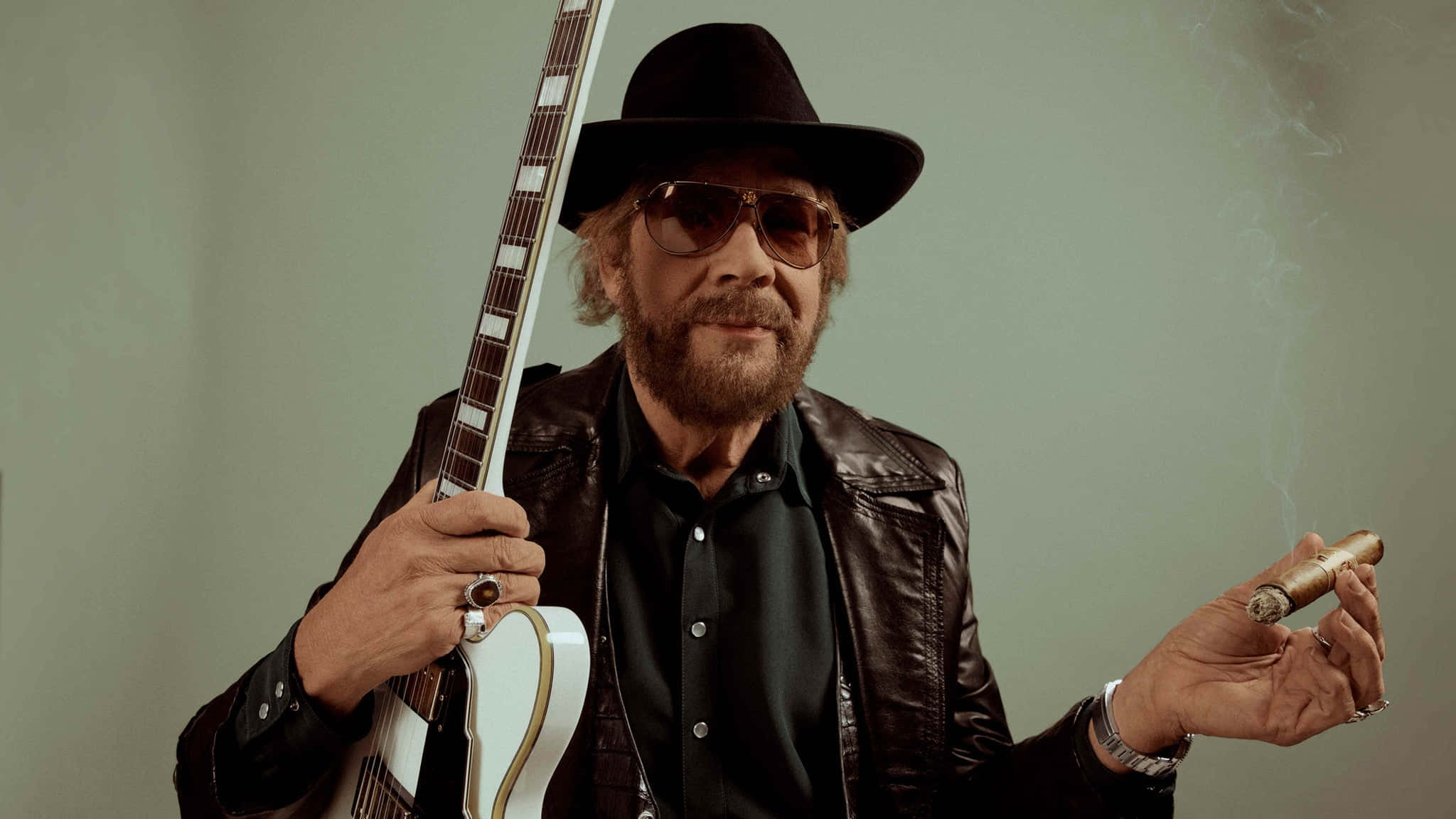 Hank Williams Jr 桌布 壁紙 — 一個留鬍鬚的男人，戴著黑色紳士帽，身穿皮夾克，手持白色電吉他，在柔和的綠色背景下抽雪茄。