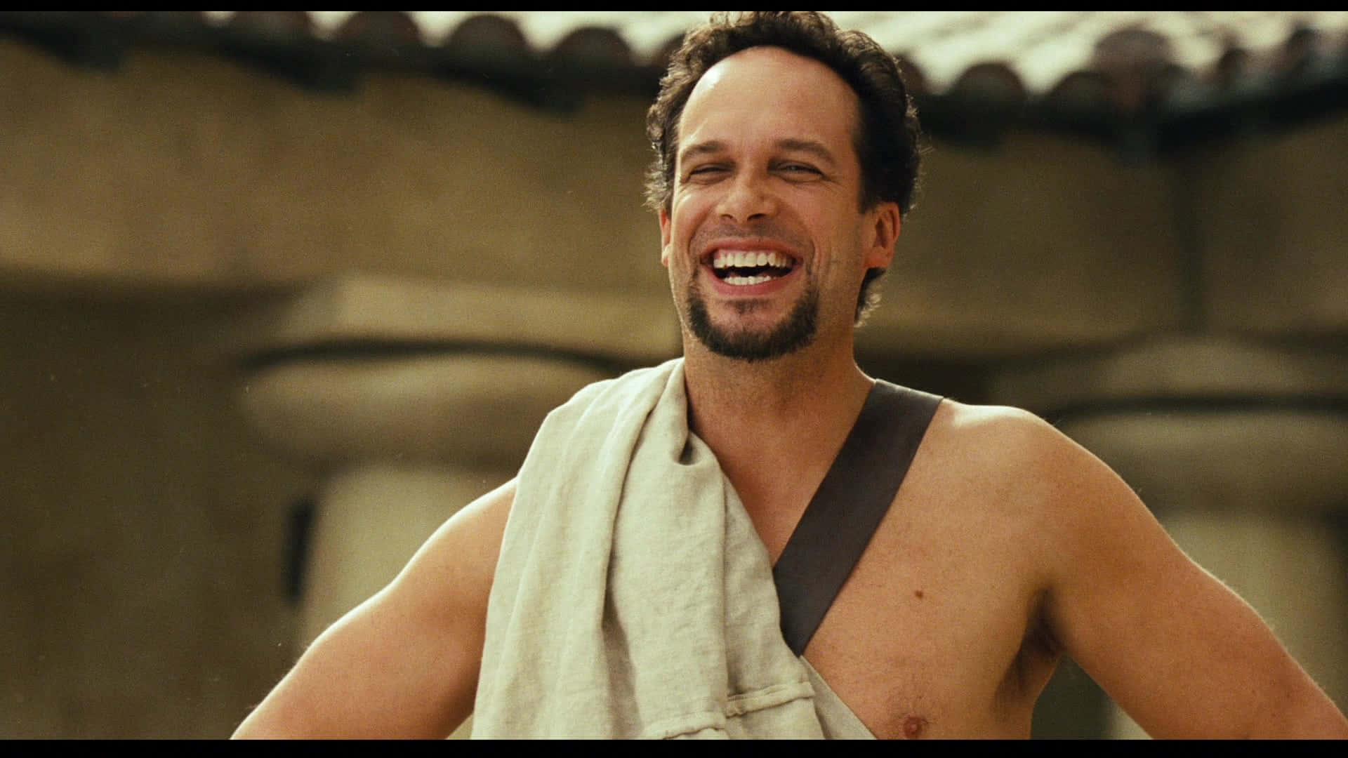 Diedrich Bader 桌布 壁紙 — 一個留鬍鬚的男人，披著毛巾，以石建築為背景，開懷大笑。