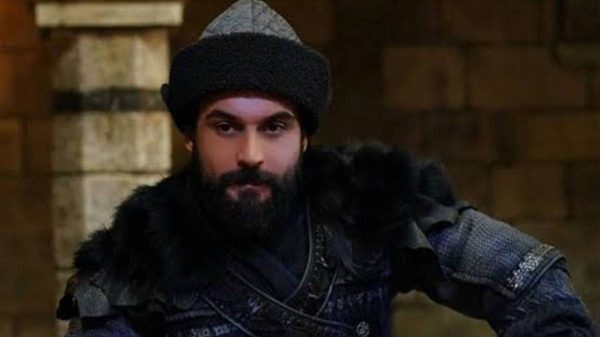 Ertugrul 桌布 壁紙 — 一個留鬍鬚的男人，身穿深色毛皮鑲邊盔甲和針織帽，在石牆背景下凝視著。