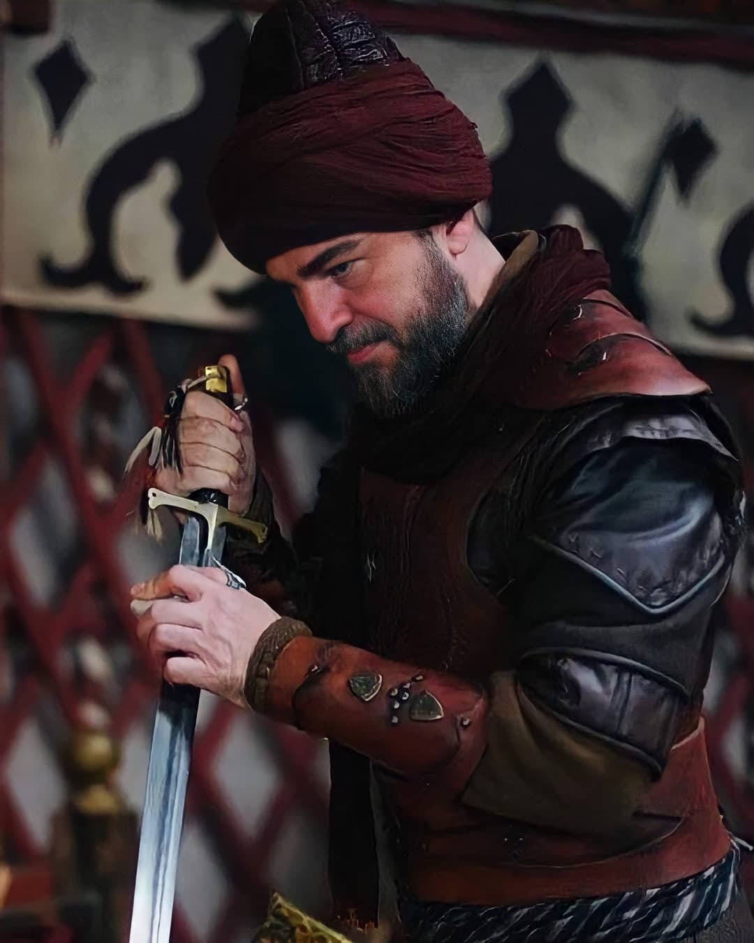 Ertugrul 桌布 壁紙 — 一個留鬍鬚的男人，身穿華麗盔甲和紅色頭巾，在圖案背景前專注地握著劍。