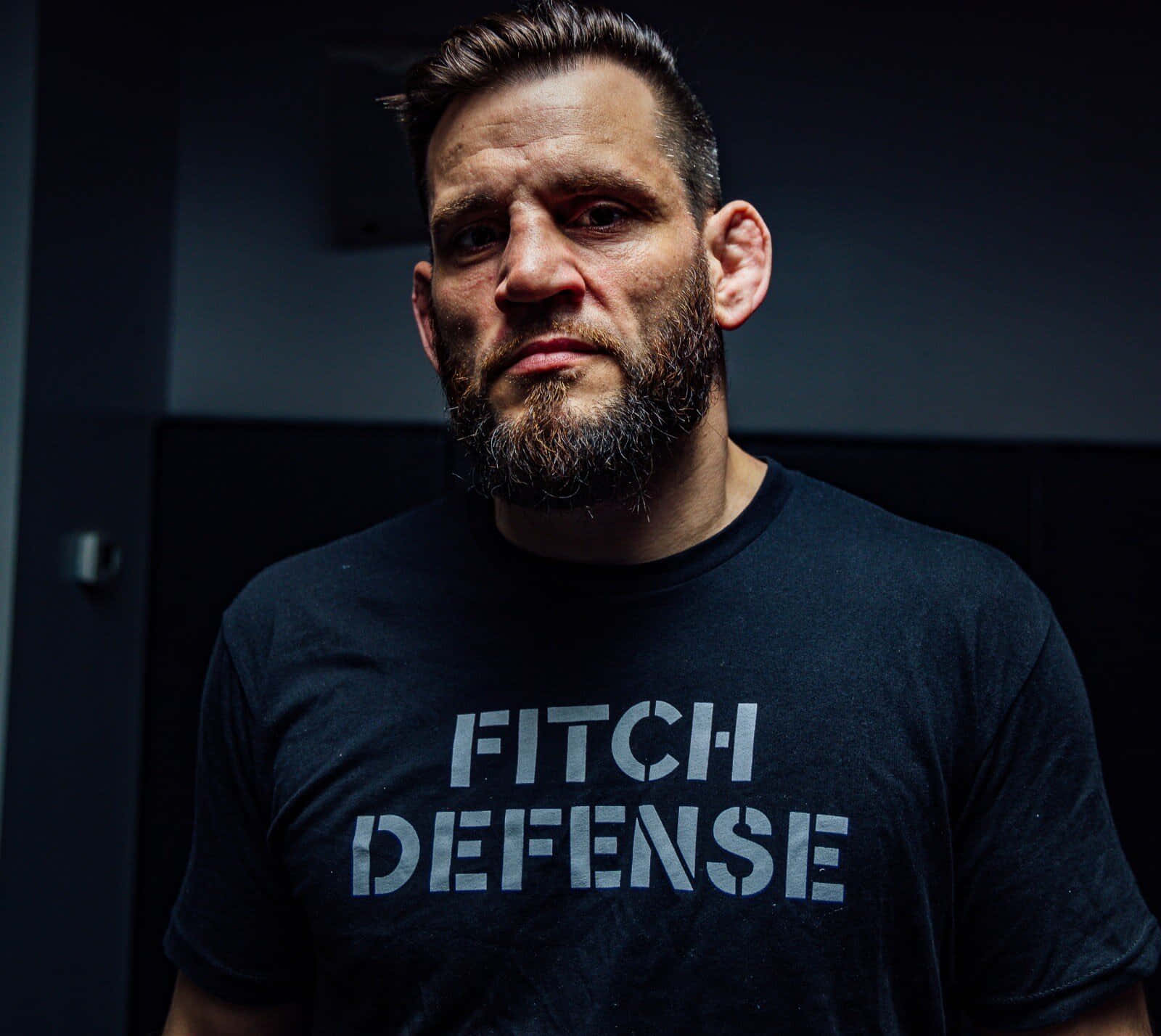 UFC 桌布 壁紙 — 一個留鬍鬚的男人，身穿黑色 'FITCH DEFENSE' T恤，在昏暗的房間裡凝視著鏡頭。