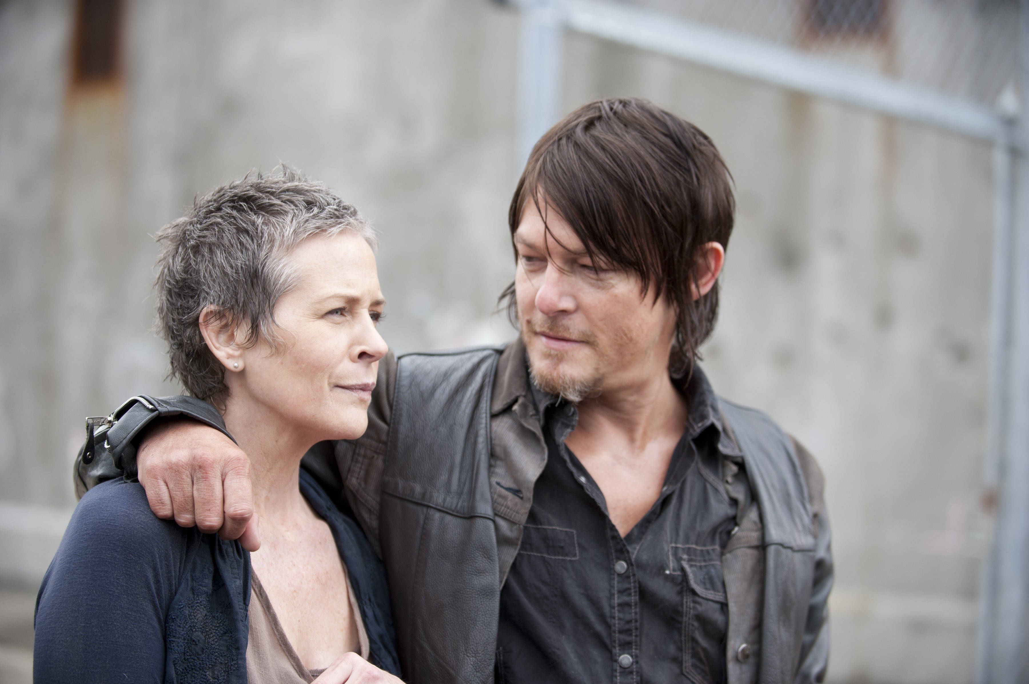 Walking Dead Daryl 桌布 壁紙 — 一個留鬍鬚的男人和一個短灰髮的女人在粗糙的城市環境中緊密地站在一起，他的手臂環繞著她的肩膀。