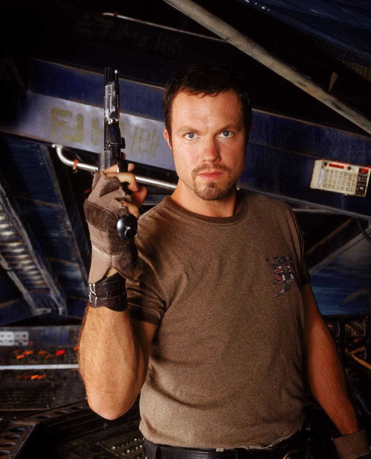 adam baldwin 桌布 壁紙 — 一個留鬍鬚的男人在昏暗的太空船駕駛艙中，手持一把雷射槍，周圍有發光的控制台和頭頂的金屬橫樑。