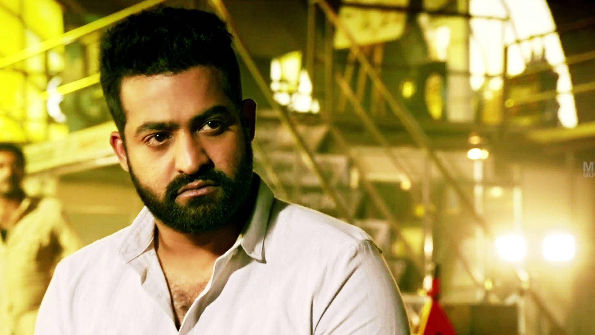 jr ntr 桌布 壁紙 — 一個留鬍鬚的男人在模糊的工業環境中，在溫暖的黃燈下專注地凝視著相機。