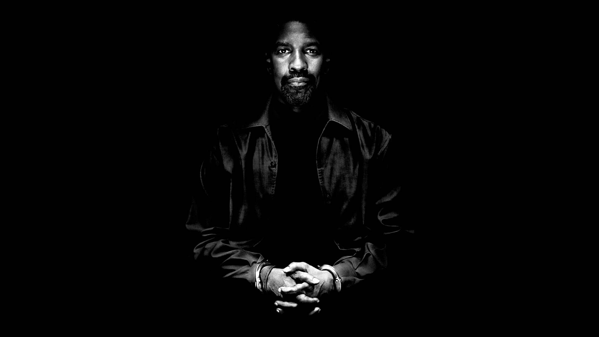 Denzel Washington 桌布 壁紙 — 一個留鬍鬚的男人在黑暗中雙手合十坐著，被戲劇性的燈光照亮，形成一幅鮮明的黑白肖像。