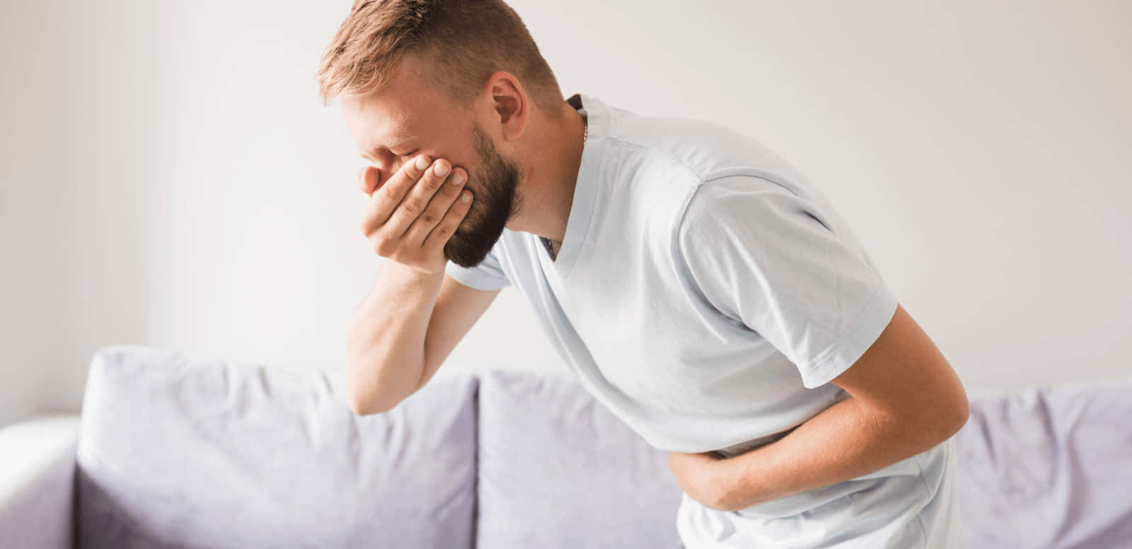 生病 桌布 壁紙 — 一個留鬍鬚的男人坐在明亮的房間裡的淺灰色沙發上，痛苦地捂著肚子。