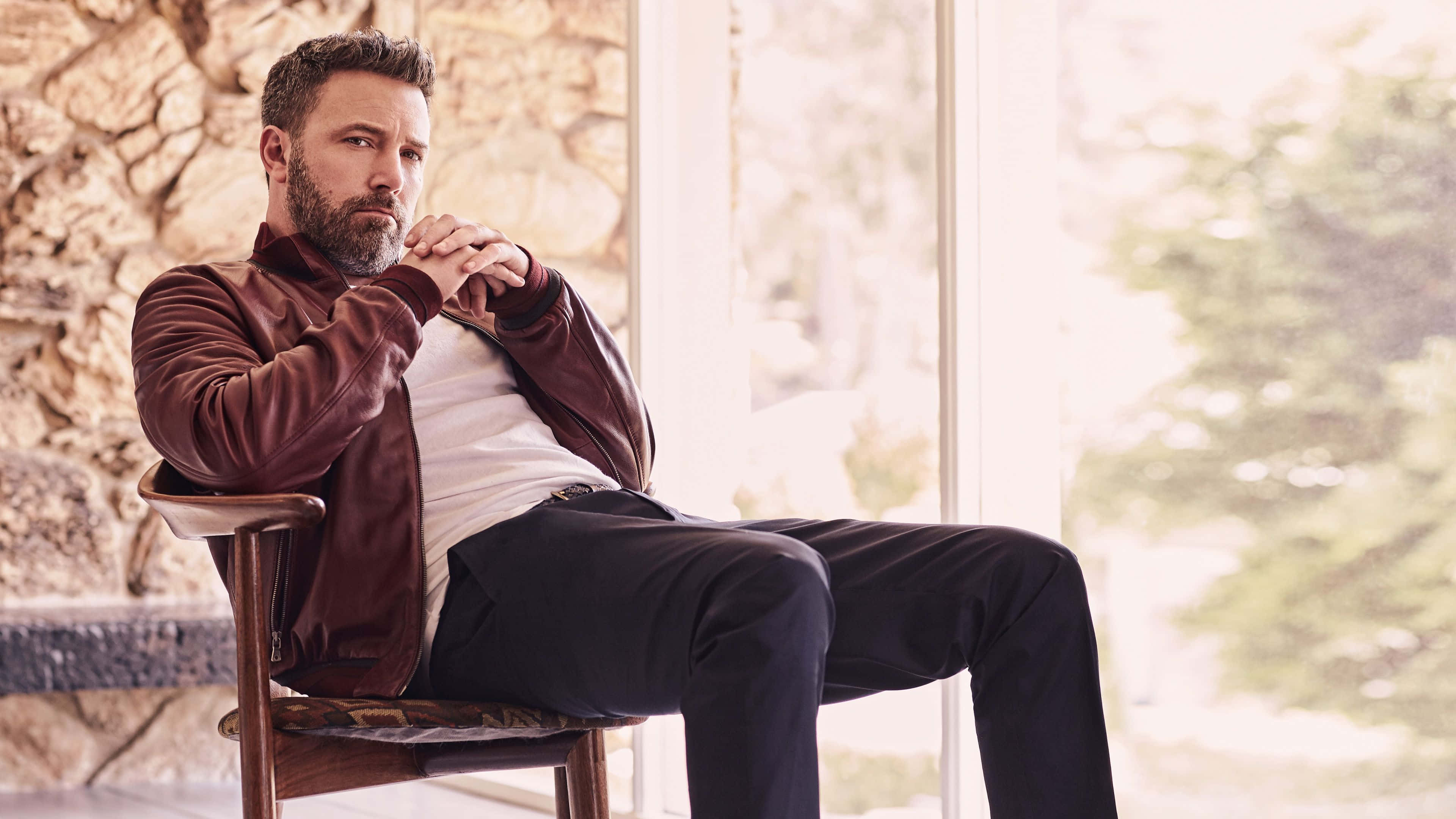 Ben Affleck 桌布 壁紙 — 一個留鬍鬚的男人坐在明亮的窗邊木椅上，背景是石牆，若有所思。