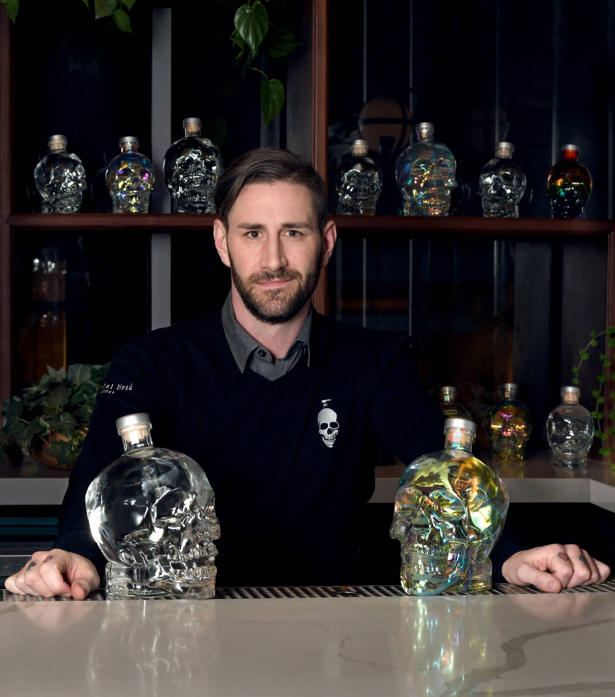 Drink 桌布 壁紙 — 一個留鬍鬚的男人坐在酒吧後面，兩旁是骷髏形狀的酒瓶，內部呈現彩虹色。