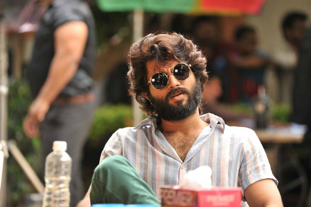 vijay devarakonda 4K 桌布 壁紙 — 一個留鬍鬚的男人戴著圓形太陽眼鏡，穿著條紋衫，隨意地坐在戶外。