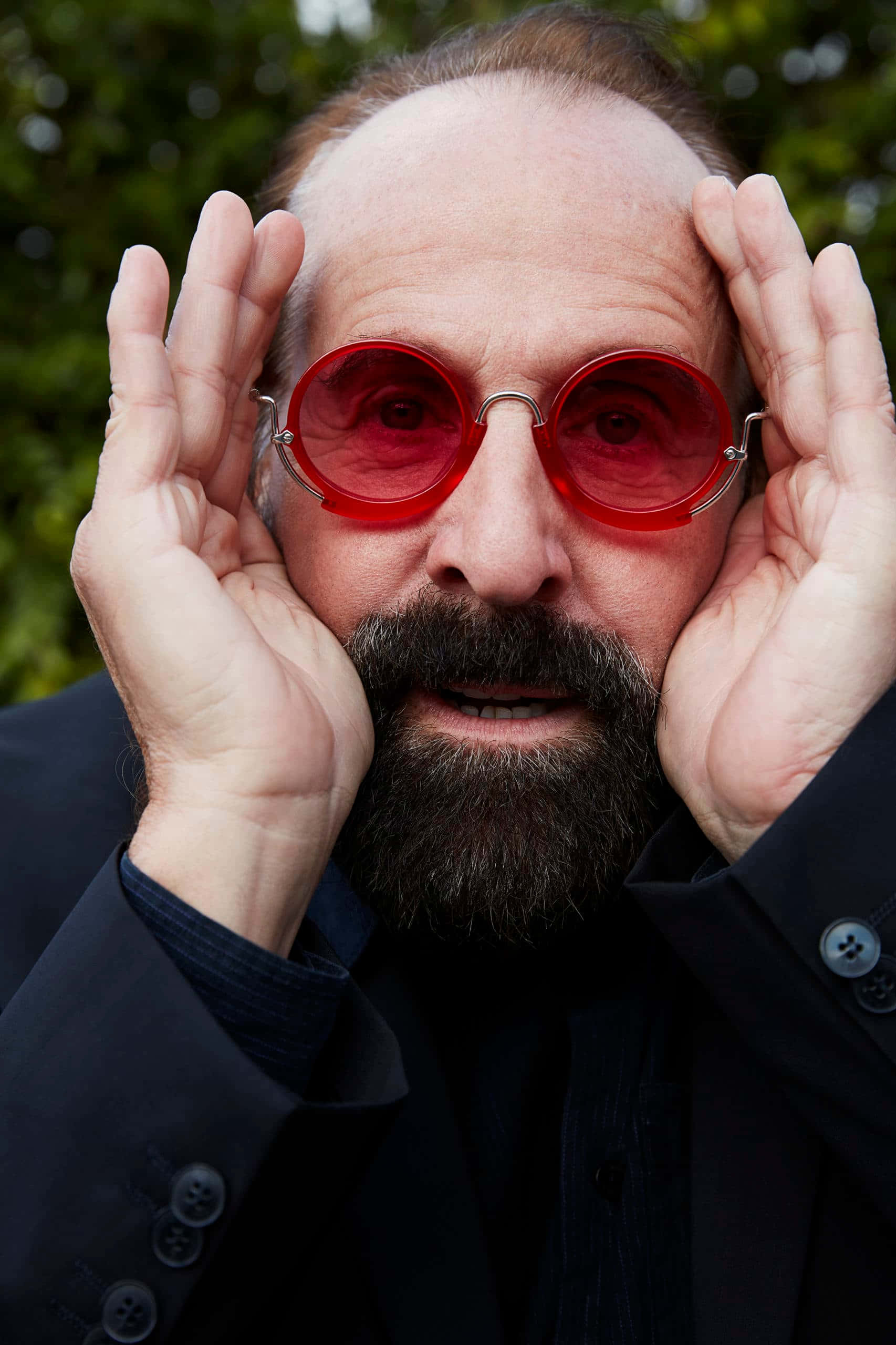 Peter Stormare 桌布 壁紙 — 一個留鬍鬚的男人戴著大膽的紅色圓形太陽眼鏡，雙手放在頭部兩側，背景是模糊的綠色。