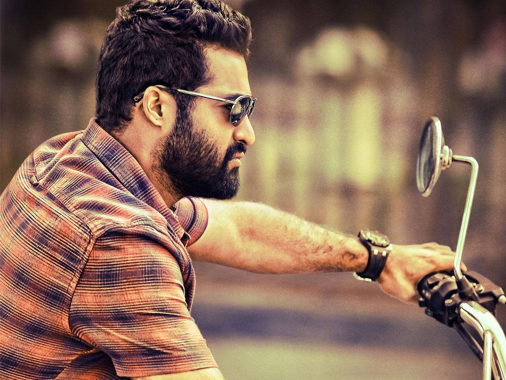 jr ntr 桌布 壁紙 — 一個留鬍鬚的男人戴著太陽眼鏡，在模糊的城市背景下，專注地握著機車把手。