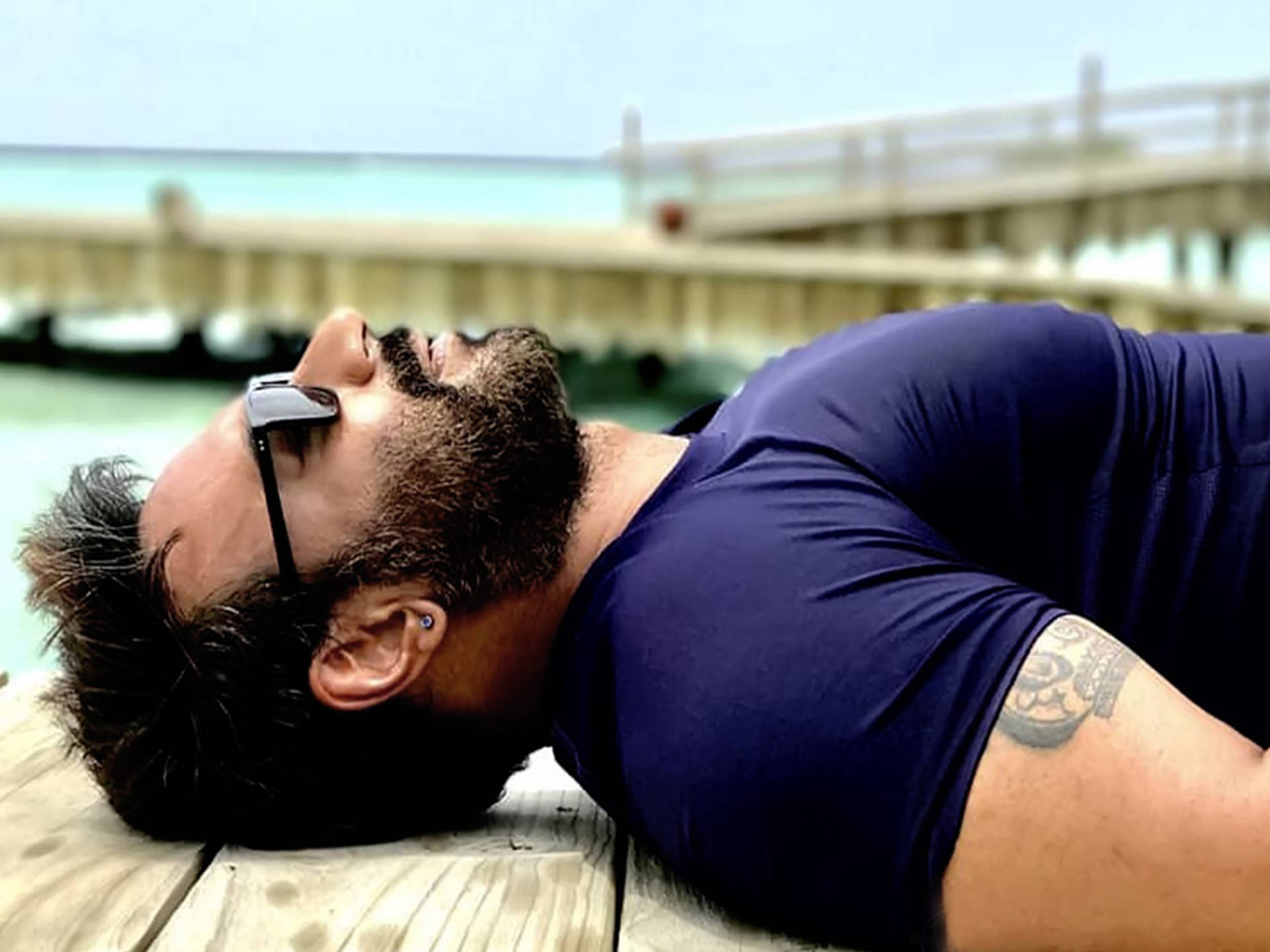 ajay devgn 桌布 壁紙 — 一個留鬍鬚的男人戴著太陽眼鏡，將頭靠在一個木製碼頭上，俯瞰著晴朗天空下的碧綠海水。