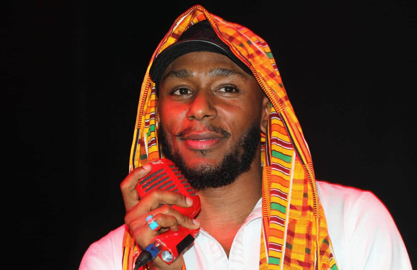 Yasiin Bey 桌布 壁紙 — 一個留鬍鬚的男人戴著彩色圖案連帽衫，在黑色背景前拿著紅色麥克風。