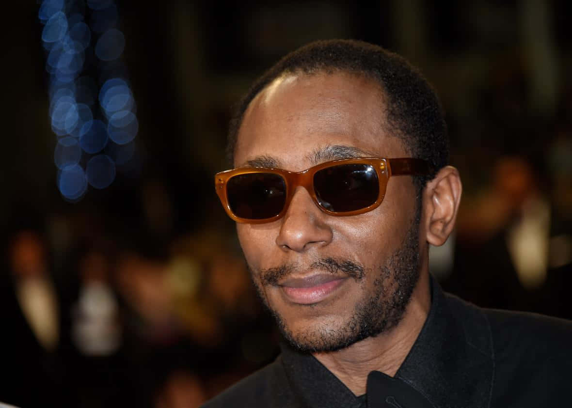 Yasiin Bey 桌布 壁紙 — 一個留鬍鬚的男人戴著時尚琥珀色太陽眼鏡，背景是模糊的藍色散景燈光。
