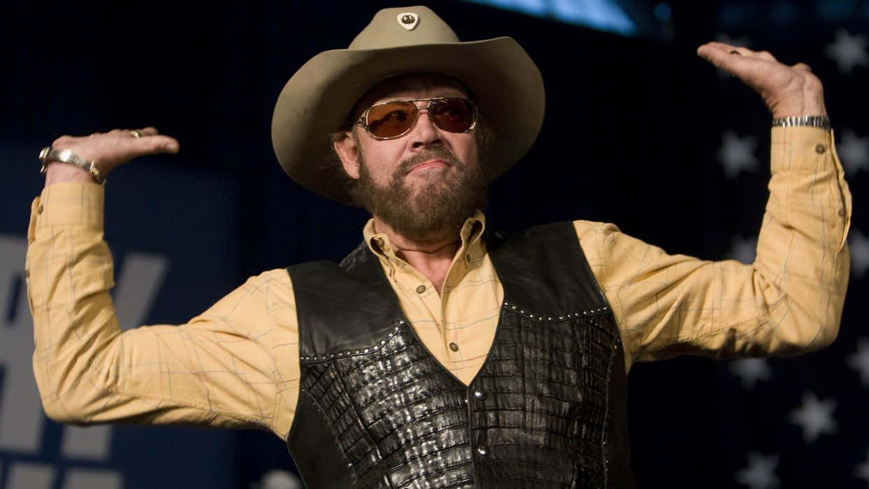 Hank Williams Jr 桌布 壁紙 — 一個留鬍鬚的男人戴著牛仔帽和太陽眼鏡，在一個有星星背景的舞台上彎曲手臂。