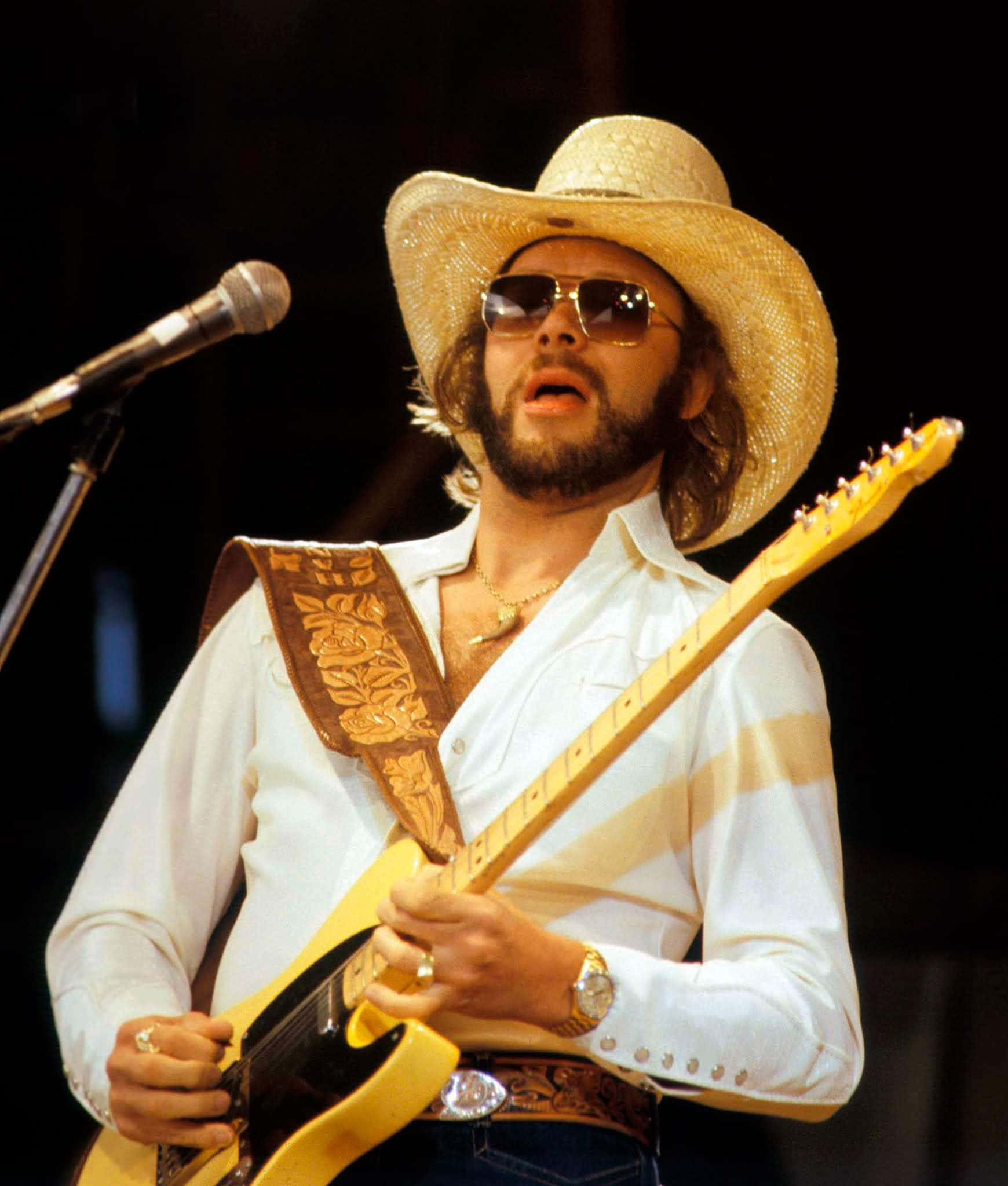 Hank Williams Jr 桌布 壁紙 — 一個留鬍鬚的男人戴著草帽和太陽眼鏡，在明亮的燈光下於舞台上熱情地彈奏一把黃色電吉他。