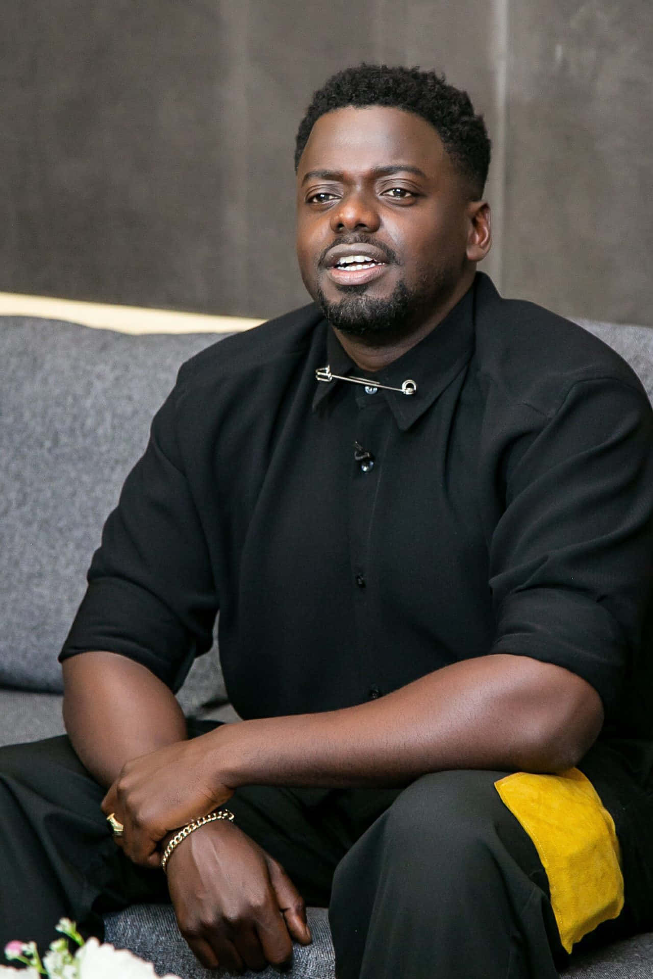 Daniel Kaluuya 桌布 壁紙 — 一個留鬍鬚的男人放鬆地坐在灰色沙發上，穿著黑色襯衫，配有銀色別針和金色飾物。