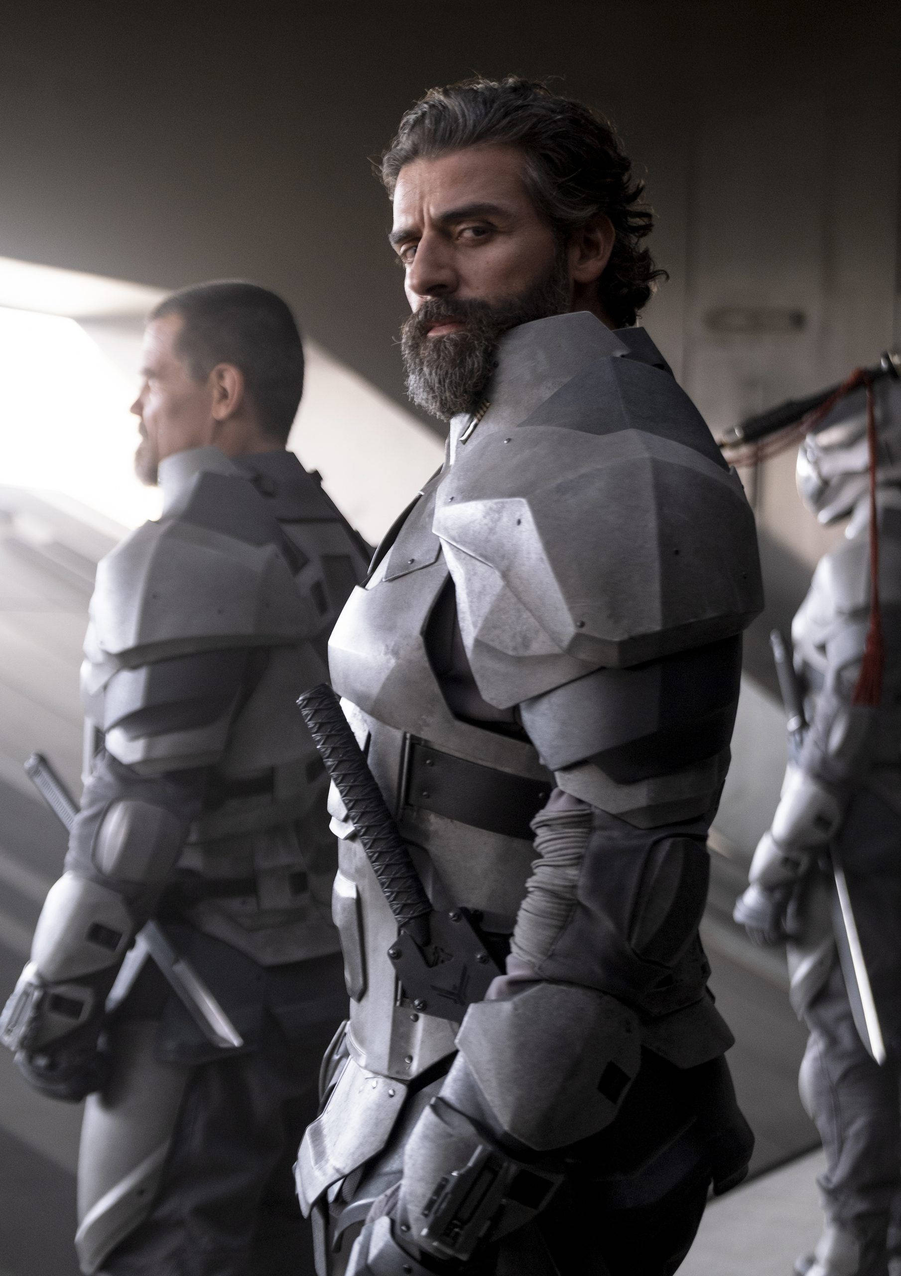 Dune Movie 桌布 壁紙 — 一個留鬍鬚的男人穿著未來風格的區塊盔甲，手持武士刀，在昏暗的走廊中被裝甲同伴簇擁。