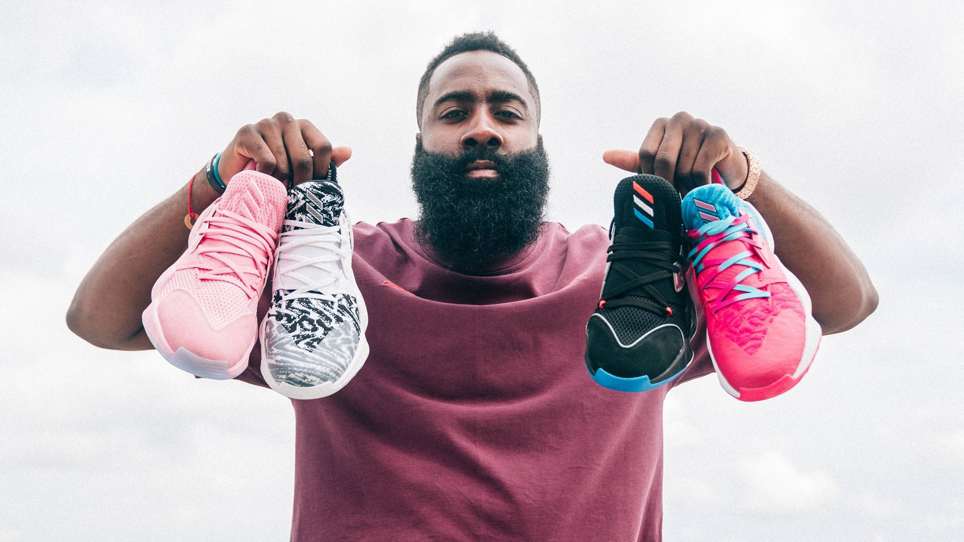James Harden 桌布 壁紙 — 一個留鬍鬚的男人穿著栗色襯衫，在陰天背景下舉起四雙色彩繽紛的運動鞋。