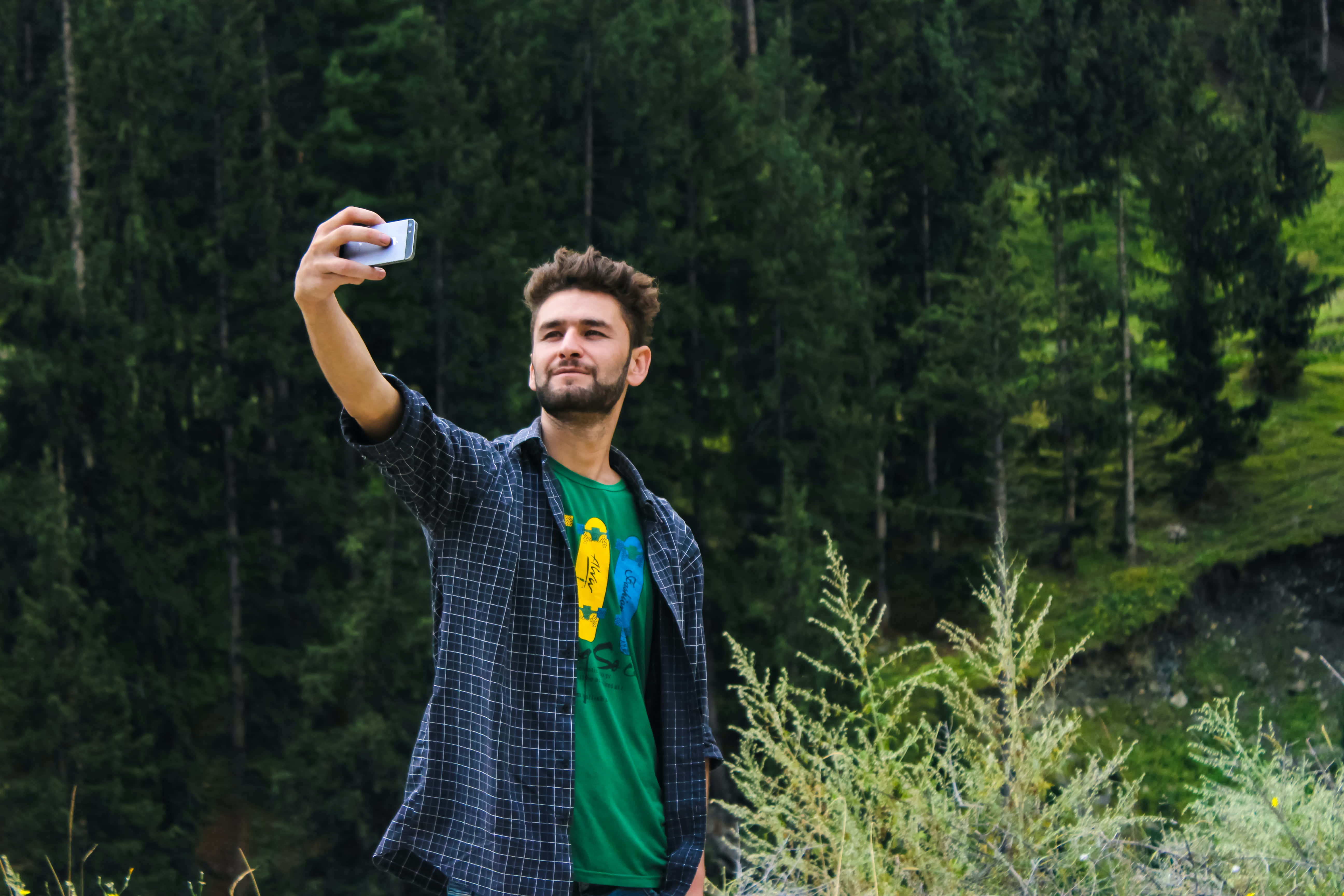 selfie 桌布 壁紙 — 一個留鬍鬚的男人穿著格紋襯衫，以茂密的綠色森林和長滿苔蘚的岩石為背景自拍。