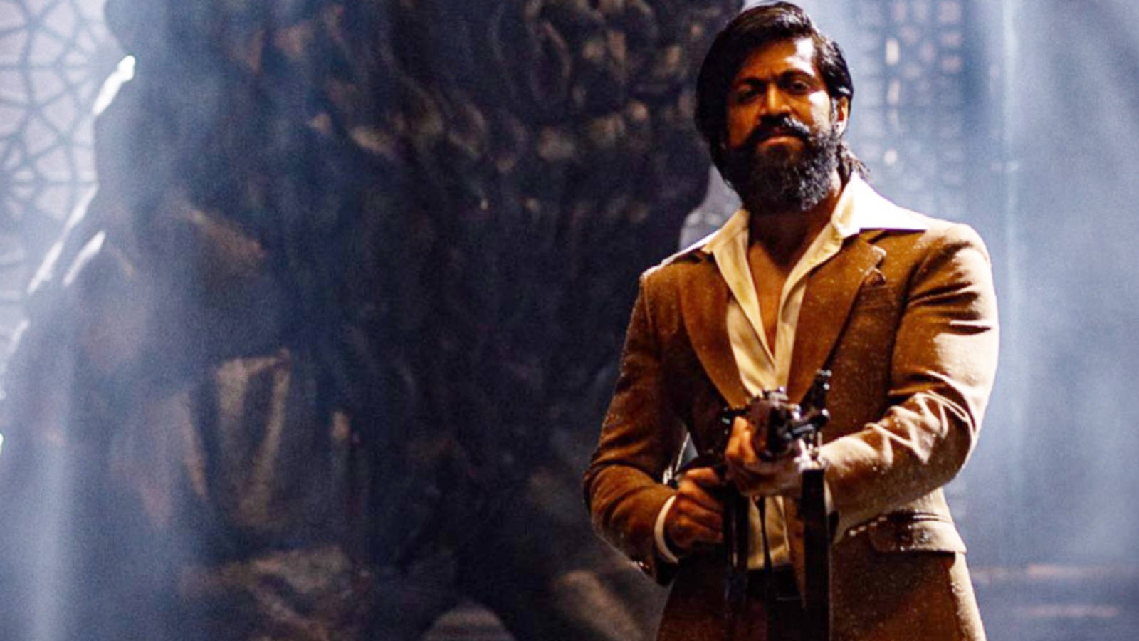 Kgf 2 桌布 壁紙 — 一個留鬍鬚的男人穿著棕色西裝，在煙霧瀰漫、裝飾華麗的石頭背景下，專注地拿著槍。