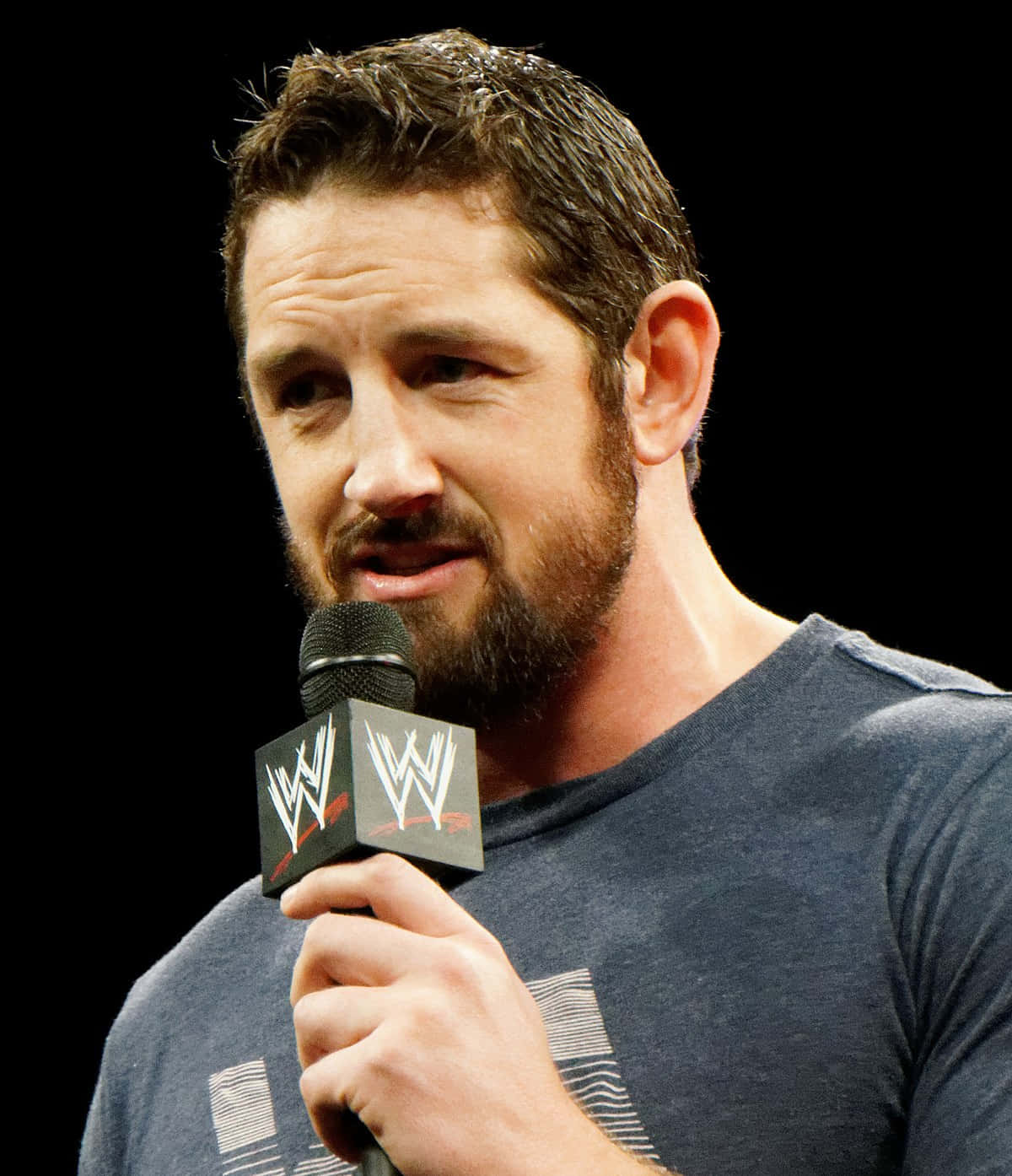 WWE Logo 桌布 壁紙 — 一個留鬍鬚的男人穿著深色T恤，在黑色背景前拿著WWE麥克風。