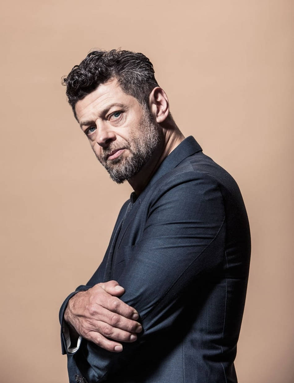 actor 桌布 壁紙 — 一個留鬍鬚的男人穿著深色西裝，自信地站在溫暖的米色背景前，雙臂交叉，直視鏡頭。
