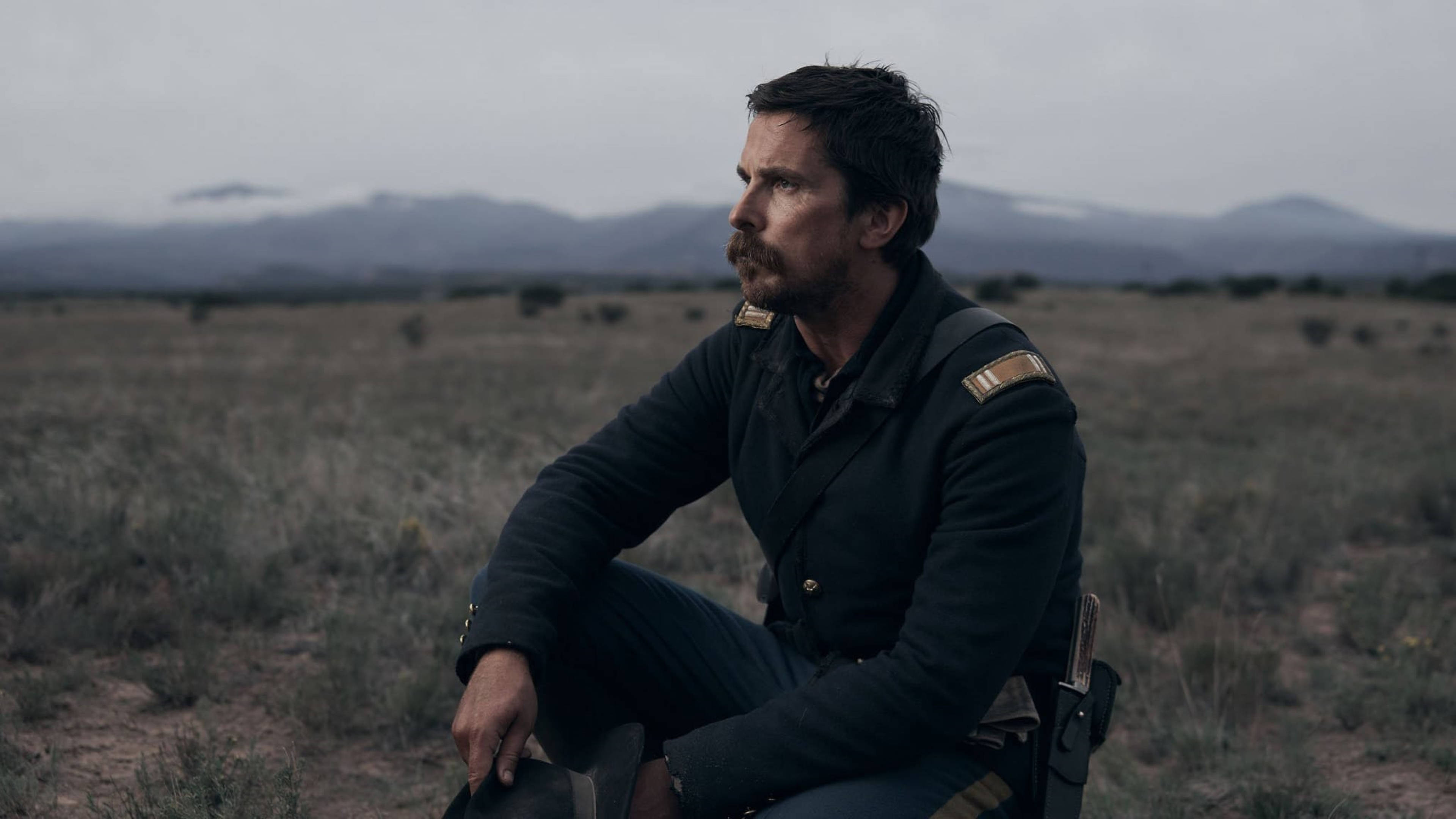 Christian Bale 桌布 壁紙 — 一個留鬍鬚的男人穿著深色軍服，獨自坐在廣闊乾燥的草地上，遠處有山脈，天空多雲。