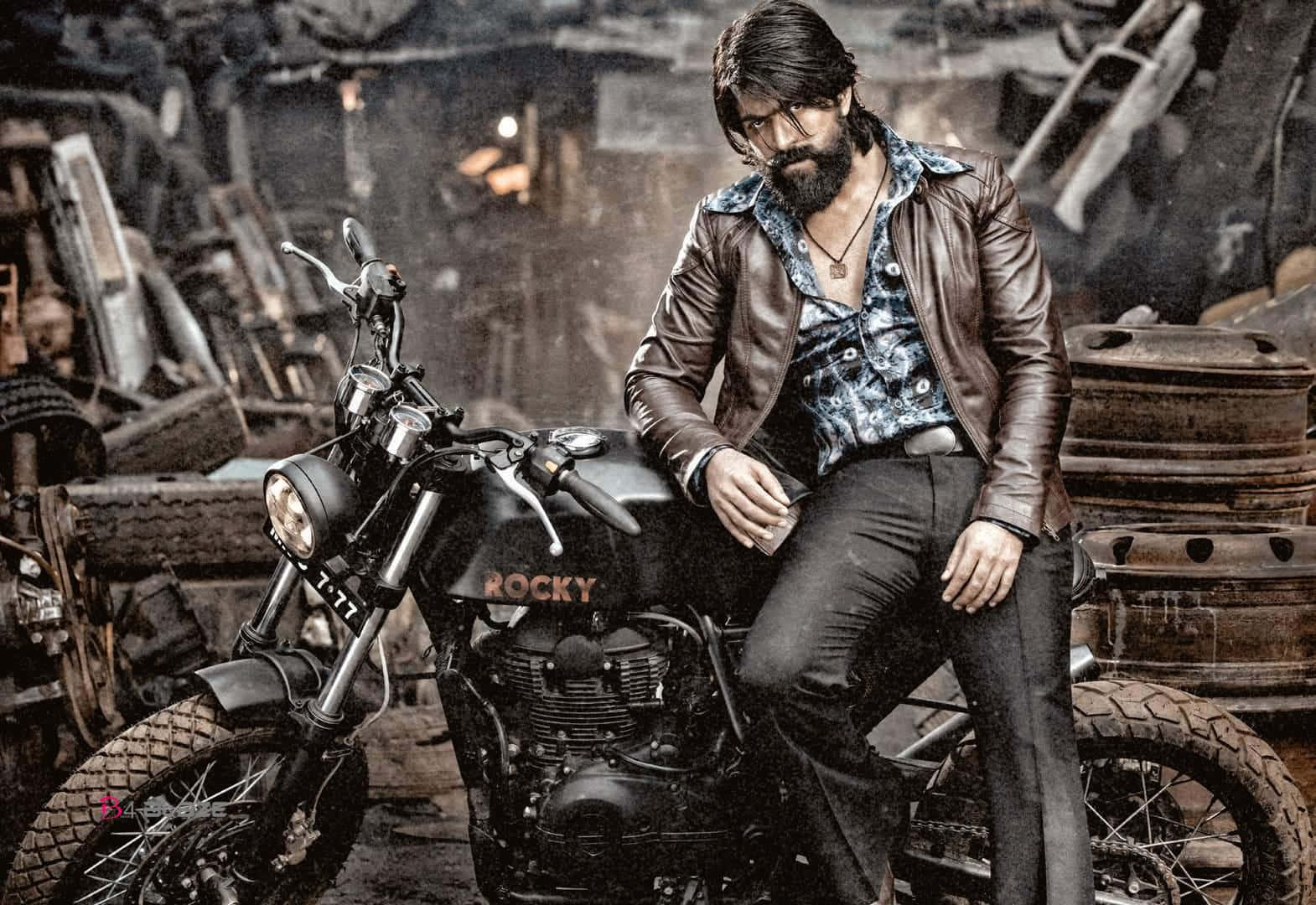 Kgf Chapter 2 桌布 壁紙 — 一個留鬍鬚的男人穿著皮夾克，自信地靠在一輛名為 'ROCKY' 的黑色摩托車上，周圍是雜亂的廢車場。